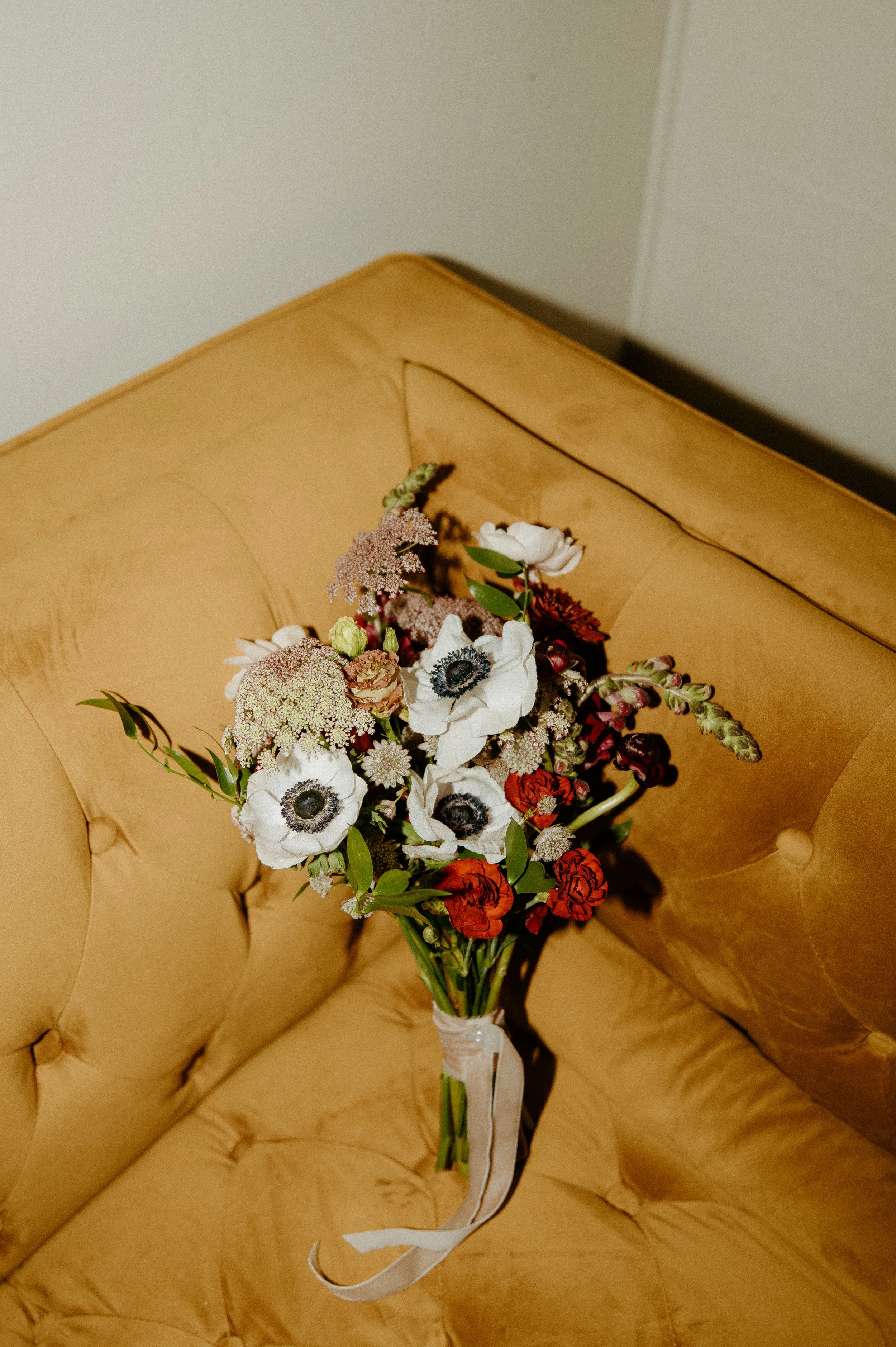 Colorful bridal bouquet with white anemones and deep red florals resting on a mustard velvet couch during a Seattle wedding