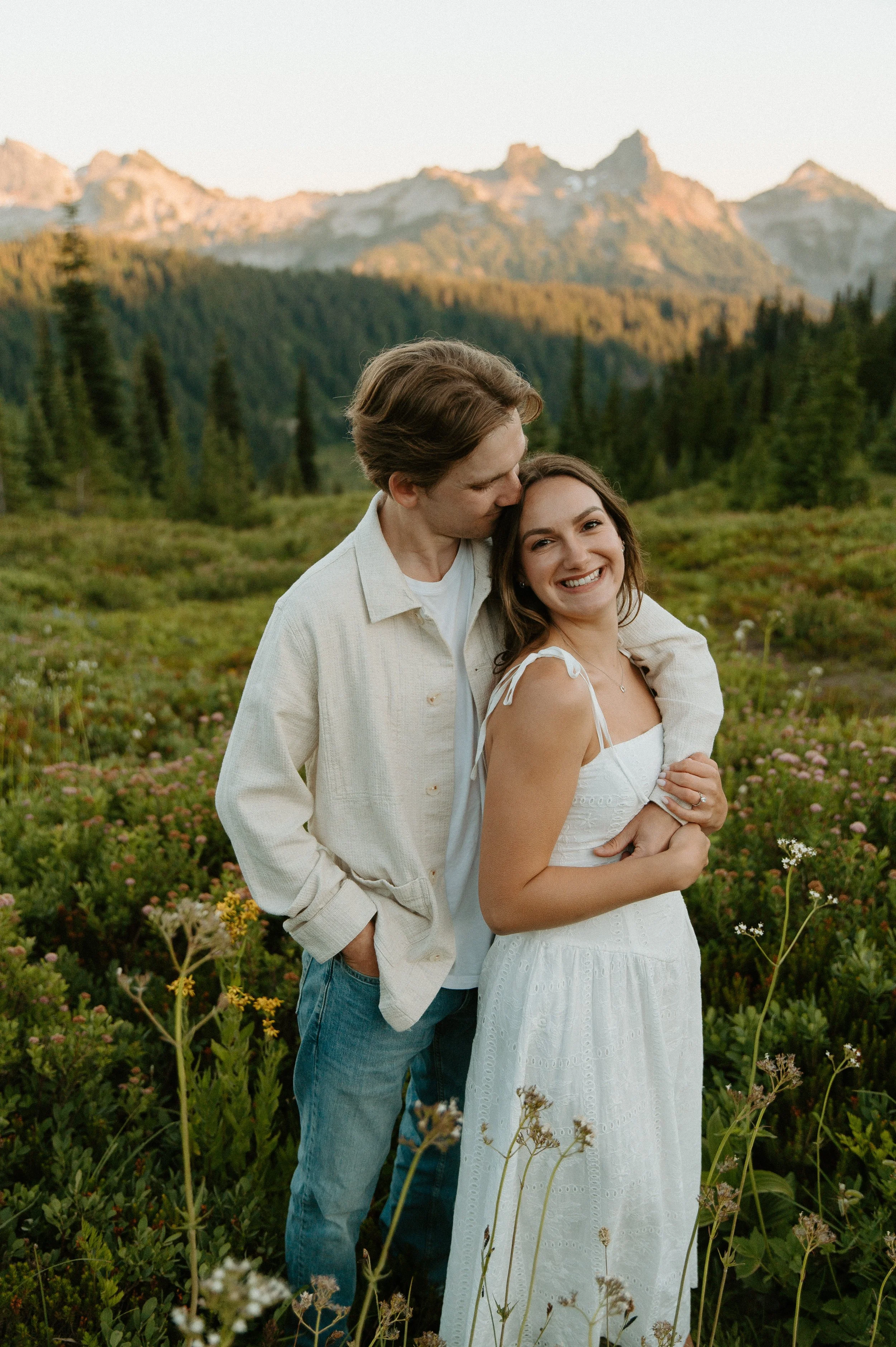 Engaged couple standing together with sunlit mountain peaks and wildflowers behind them.