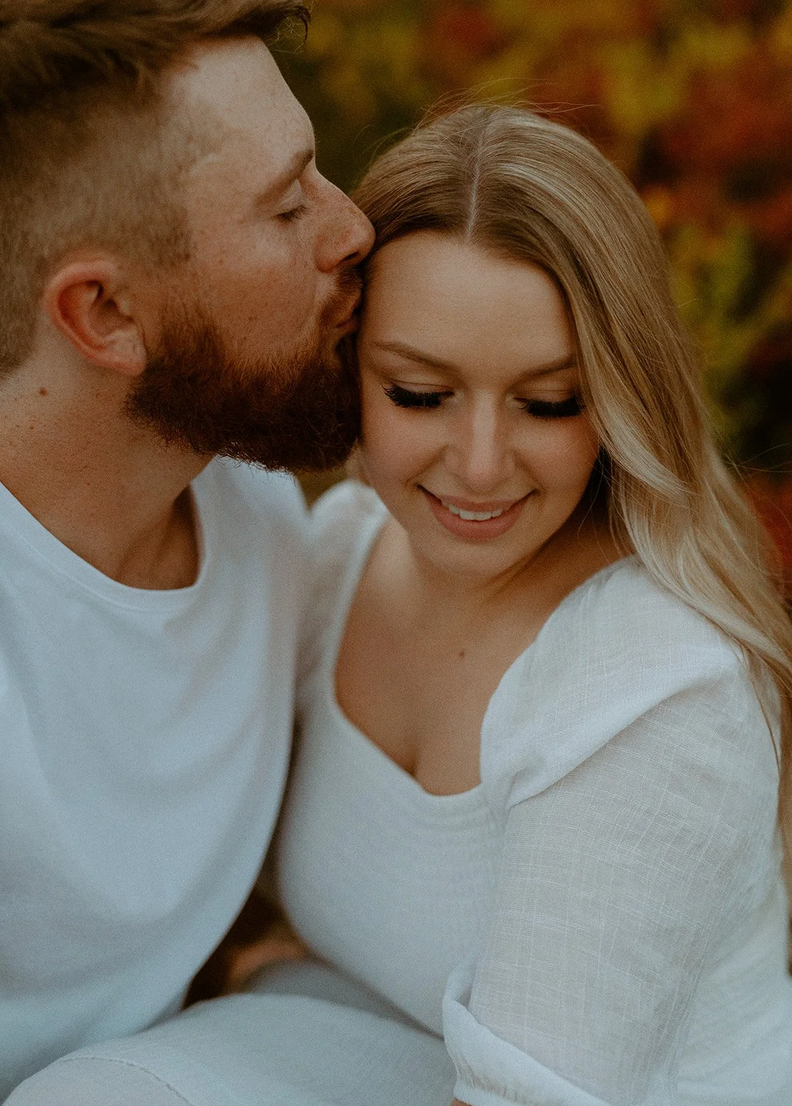 Close-up of couple embracing, groom kissing bride’s forehead with warm autumn tones in the background