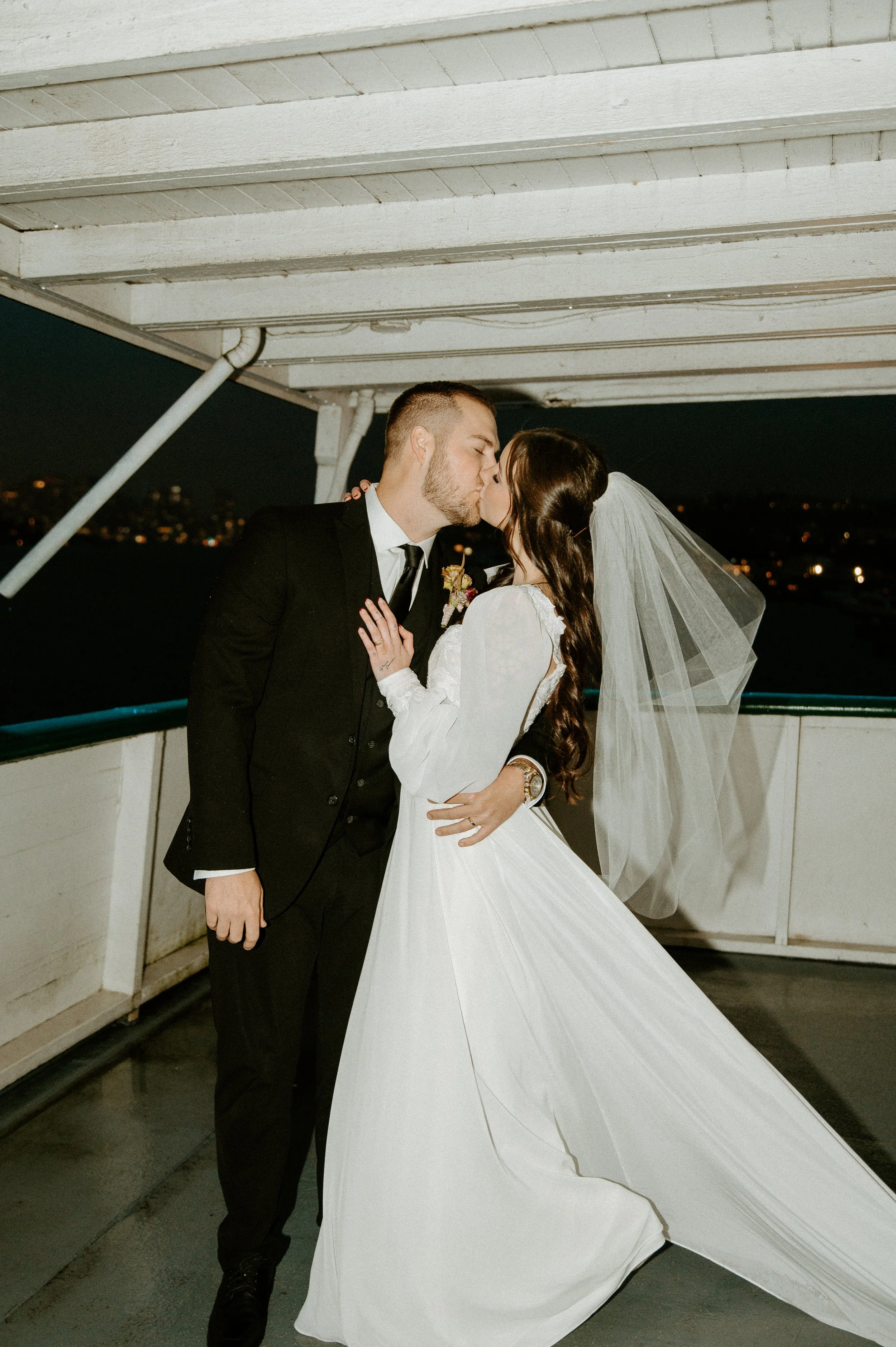 Bride and groom kissing on the deck of a boat at night with city lights in the background during a Seattle wedding reception