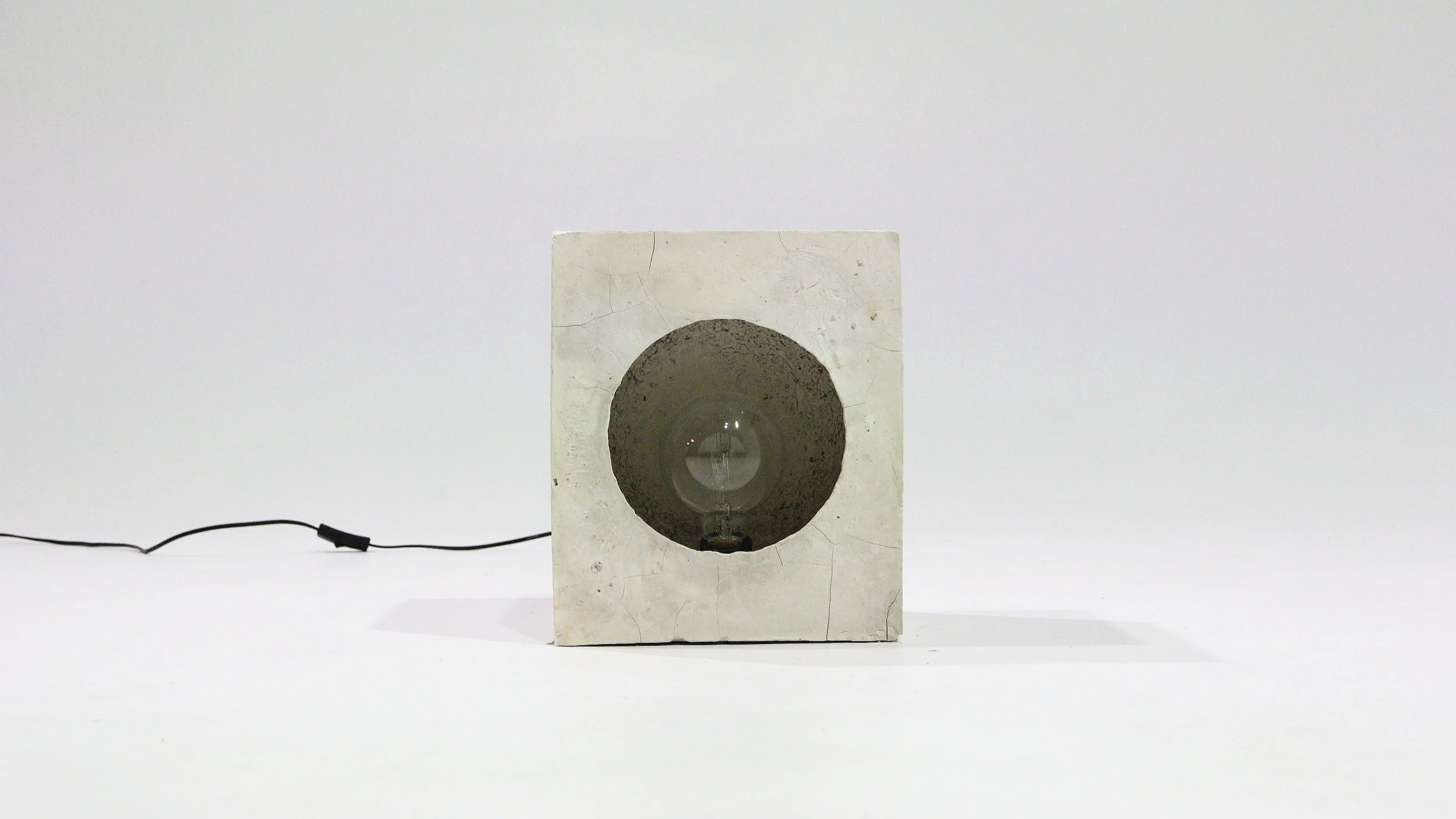 A cubic concrete speaker with a circular opening in the front, showing a glass bulb inside, connected to a black wire on a white surface with a white gradient background.