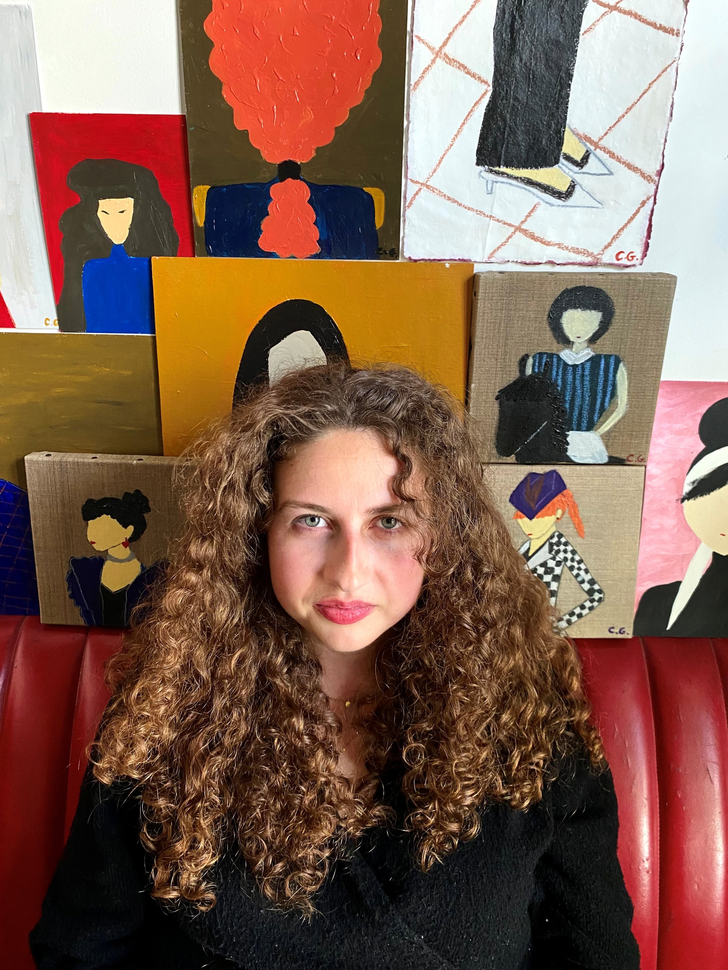 Une jeune femme aux cheveux bouclés bruns foncés pose devant plusieurs tableaux de style abstrait et illustratif, assise sur un canapé en cuir rouge.