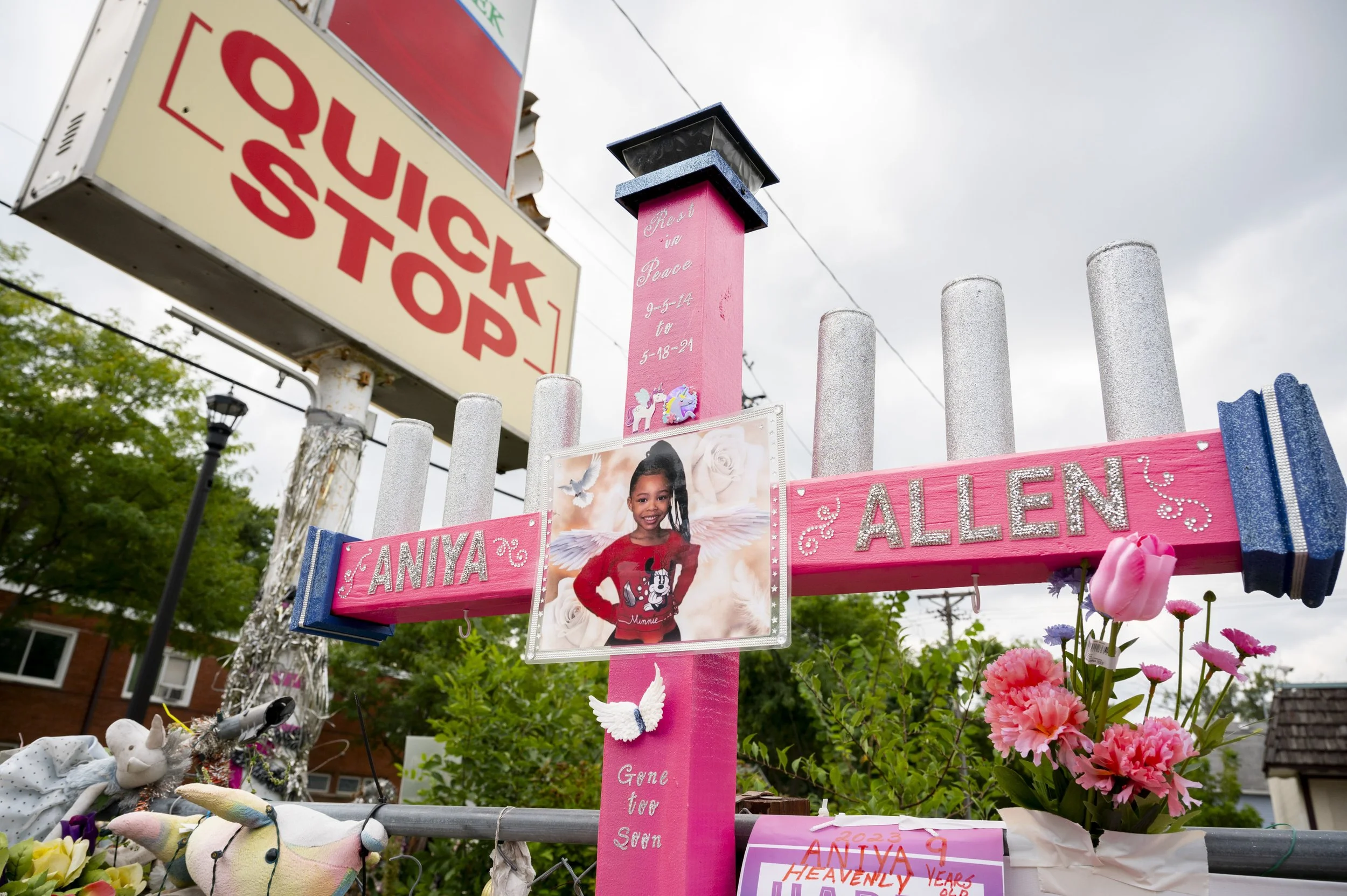 A makeshift memorial with pink and silver decorations, flowers, and a photo of a young girl named Aniya, honoring her memory with engraved messages. In the background, there's a sign that says 'Quick Stop' and a cloudy sky.