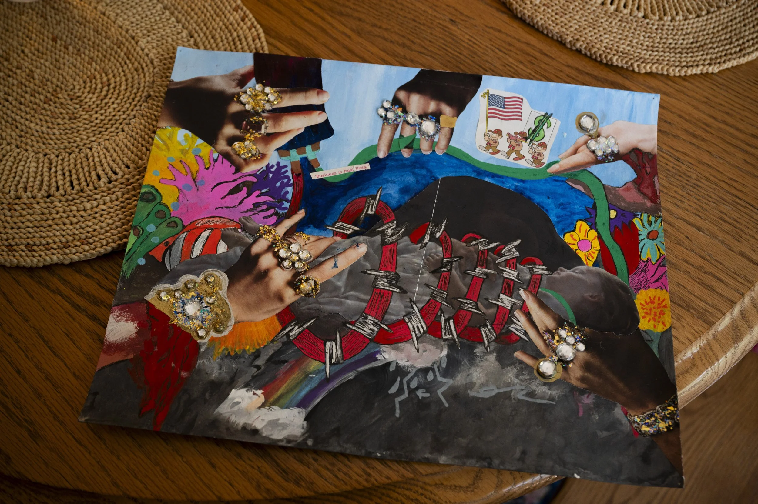 Colorful artwork of hands wearing large gem-encrusted rings and bracelets, pointing at a painted scene with American and political flags, a tree, a mountain, and a rainbow, on a wooden table.