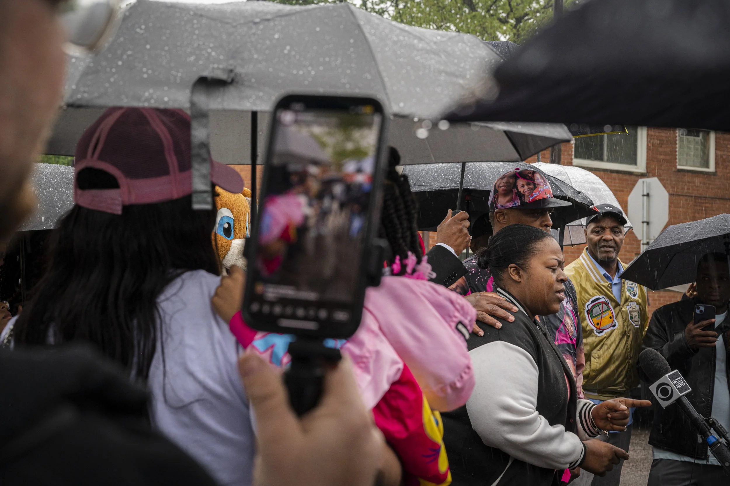 People, including women and a man in a yellow jacket, standing outside in the rain under umbrellas. One woman is speaking into a microphone, with others listening nearby. Someone is taking a photo or video with a smartphone.