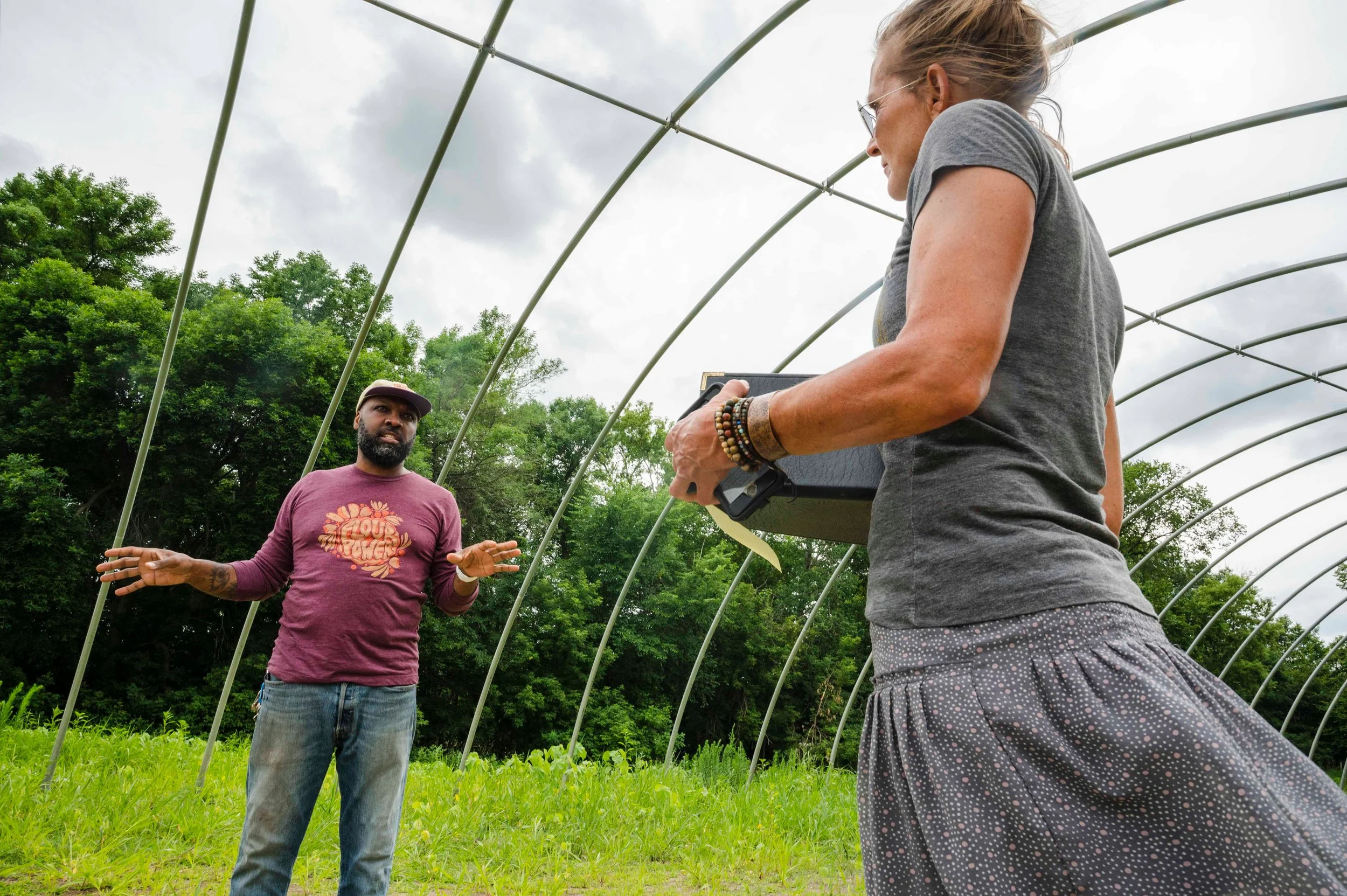 A woman being interviewed by a man outdoors in a green field with a metal arch structure overhead, trees in the background, and cloudy sky.