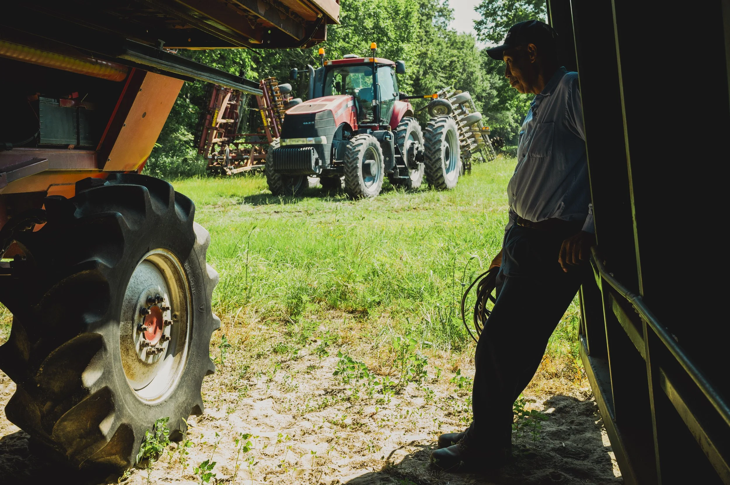 A man stands inside a barn or shed, looking at the ground, with a large red tractor outside in a grassy and wooded area.