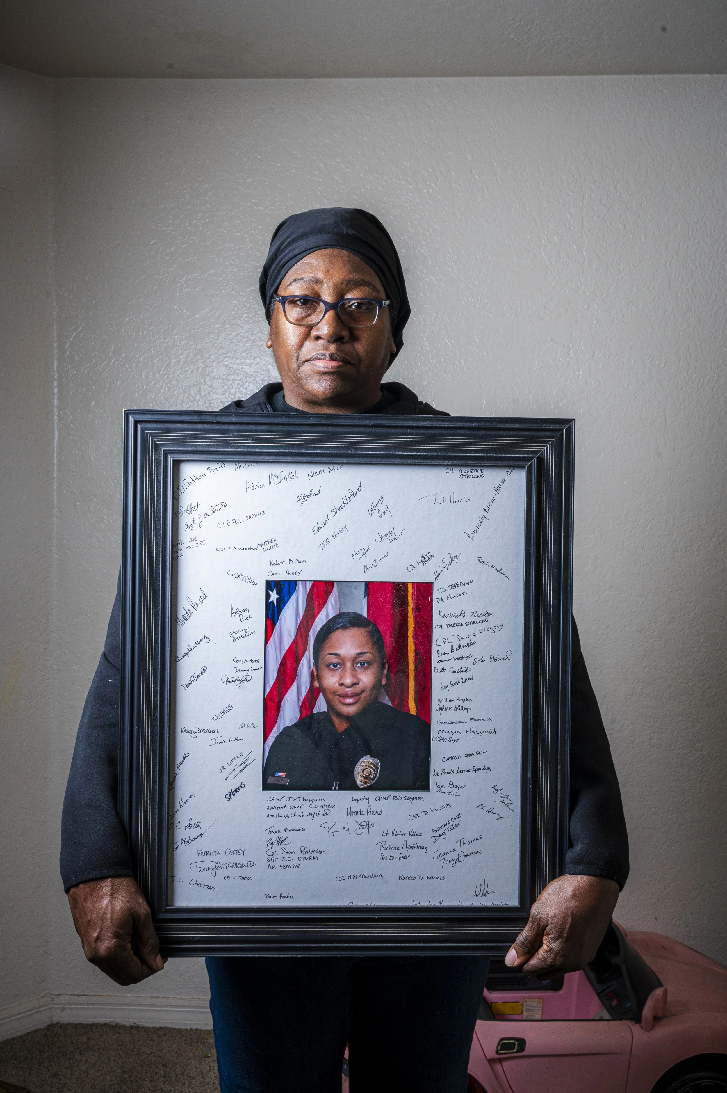 A woman holding a framed photograph of a young female police officer in front of a wall and a pink toy car on the floor. The picture frame contains a photograph of the officer with an American flag background, and signatures around it.