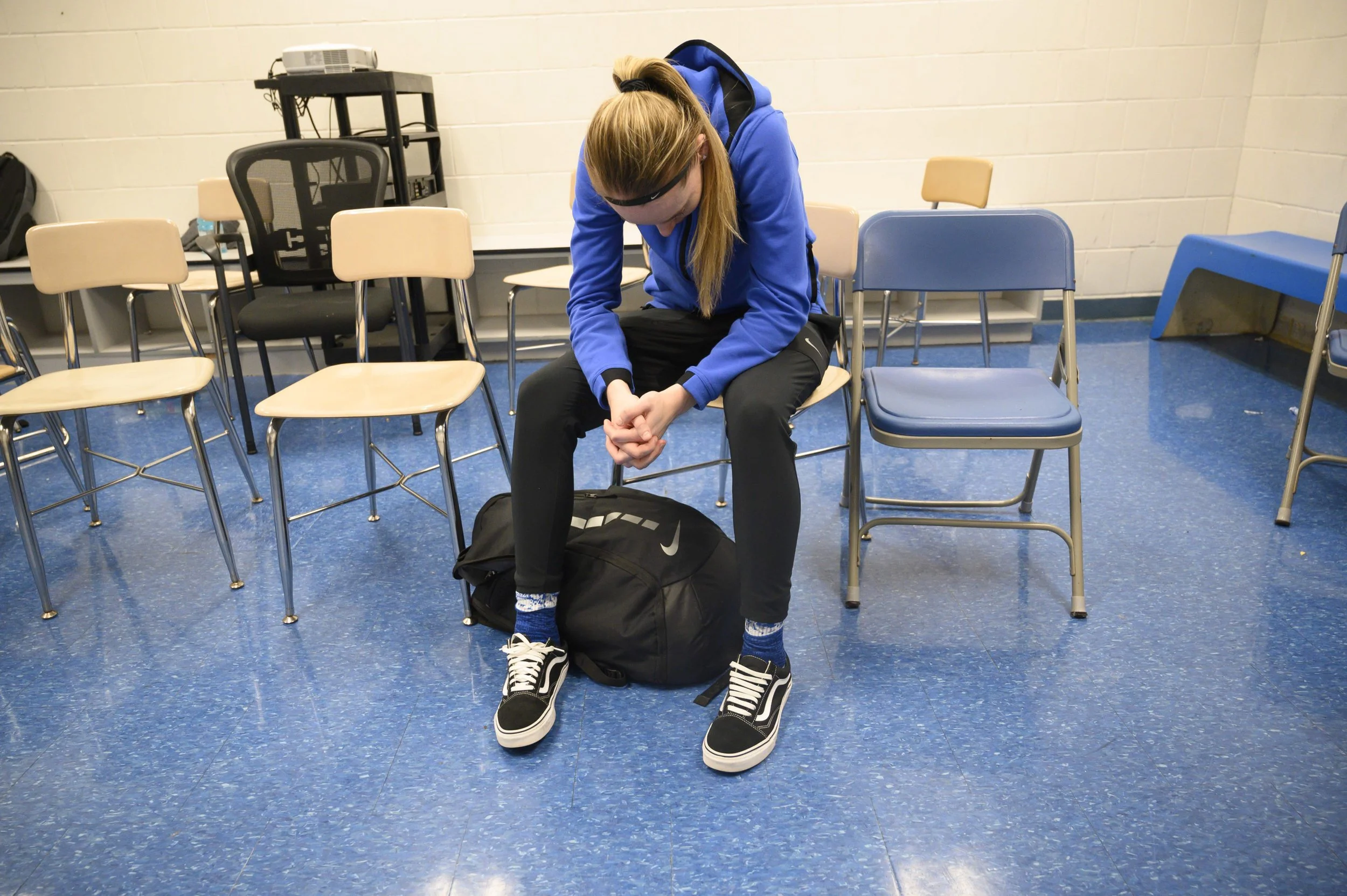 A teenage girl with red hair, wearing sunglasses, a blue hoodie, black pants, and Vans shoes, sits on a chair with her head down and hands clasped, in a room with blue tiled floor and rows of beige and blue chairs.