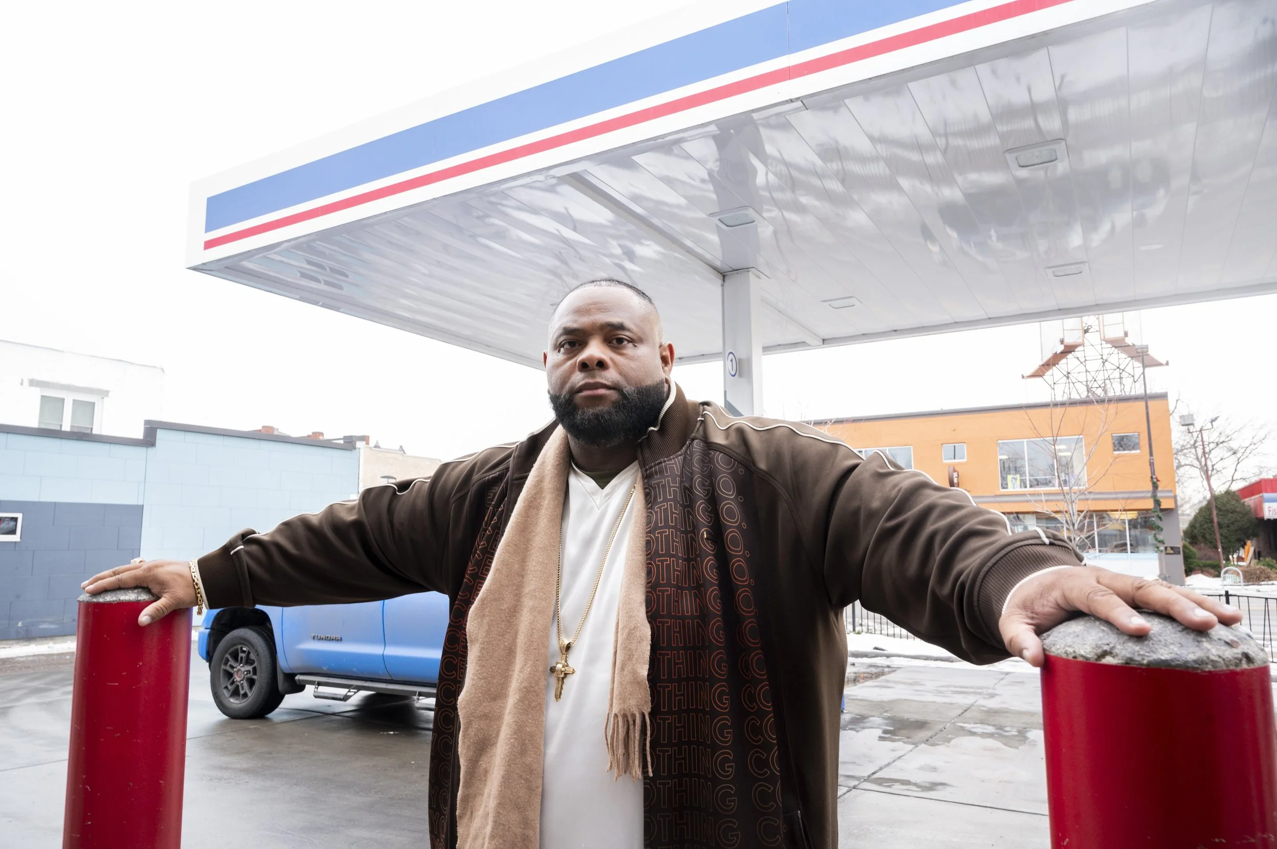 A man with a beard and short hair outdoors at a gas station, stretching his arms across red post barriers with a blue vehicle and a building in the background.