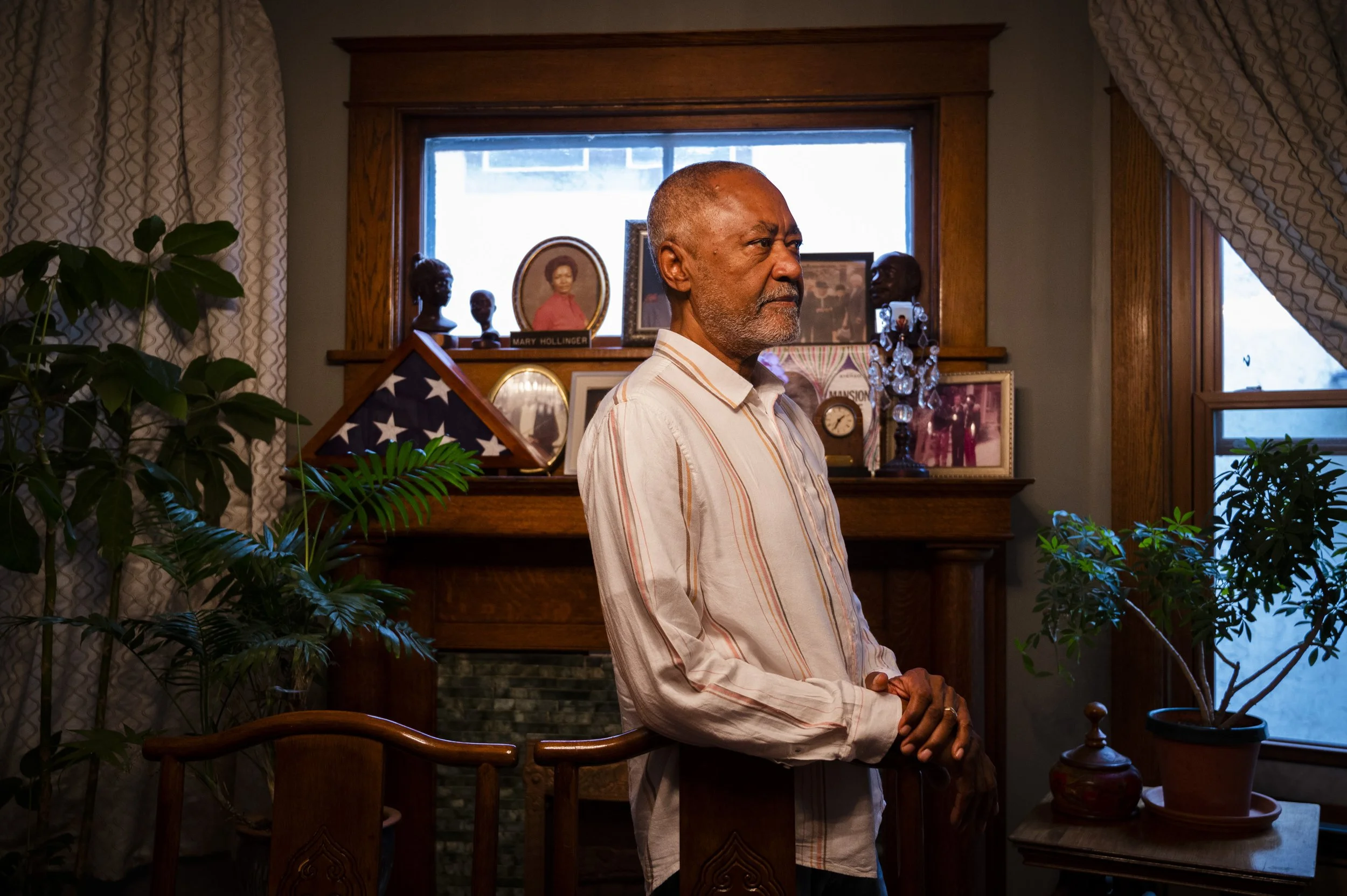 An older man with gray hair and a beard stands in a living room, facing to the right with a serious expression. He wears a light-colored striped shirt and holds a wooden cane in his right hand. The room features wooden window frames, a bookshelf with