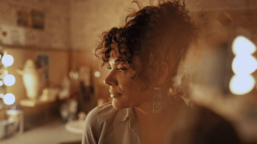 A woman with curly hair and earrings sitting in a warmly lit room.