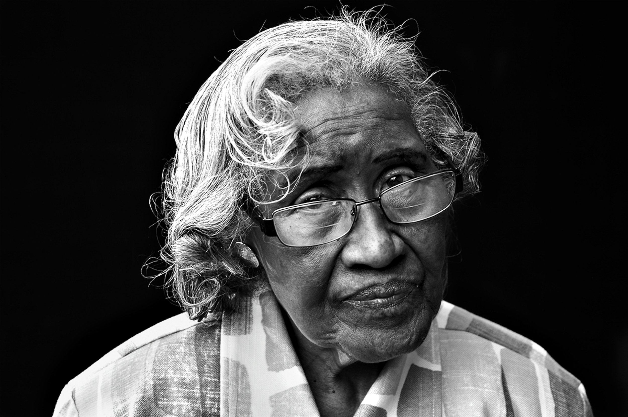 A black and white close-up portrait of an elderly woman with gray curly hair, wearing glasses and a patterned shirt, against a dark background.