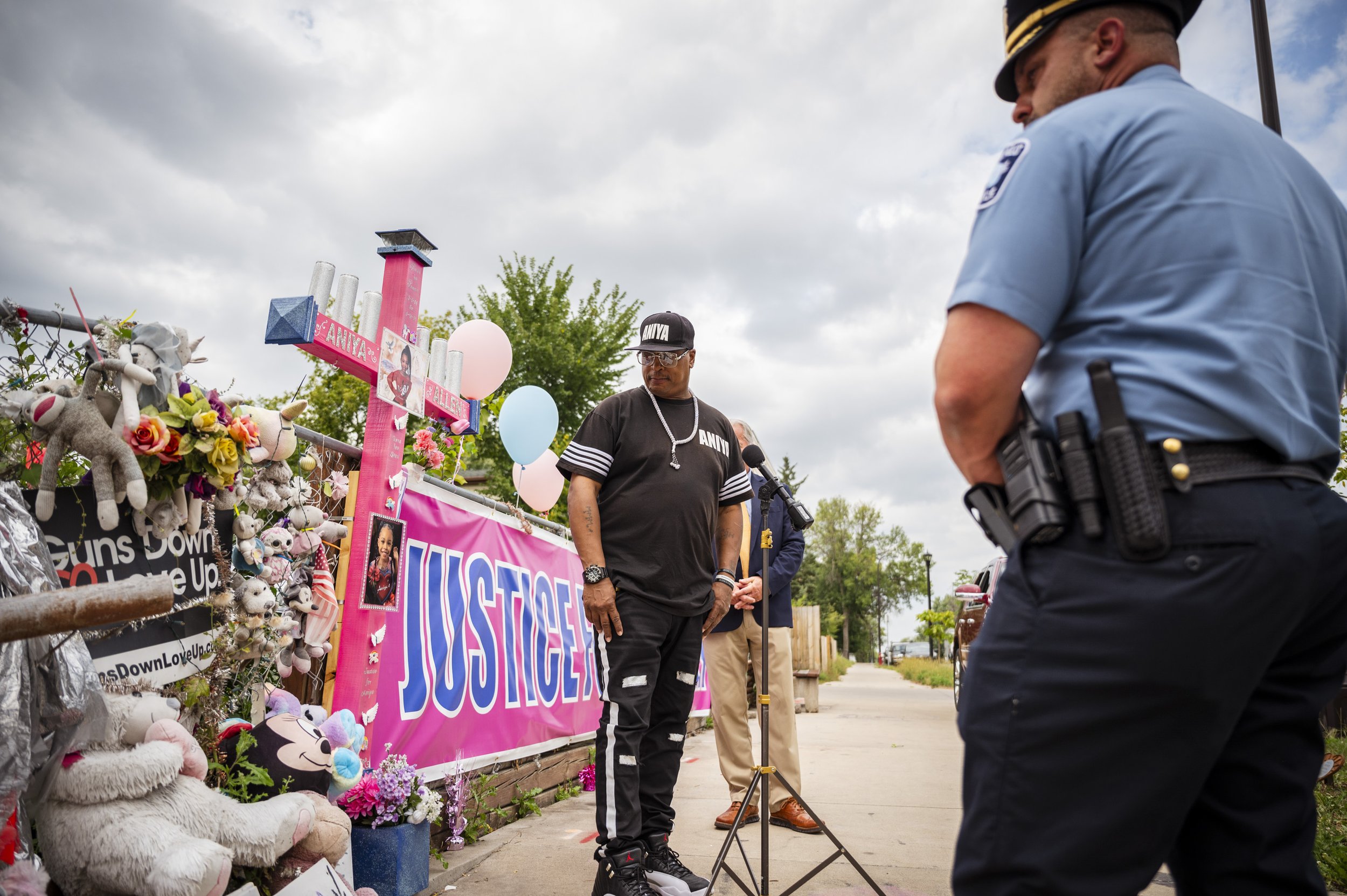 A memorial with flowers, stuffed animals, balloons, and signs, including a prominent pink sign reading 'JUSTICE.' There are two men in the scene: one man wearing a black 'ANAYA' T-shirt, black pants, and a cap standing near the memorial, and a police
