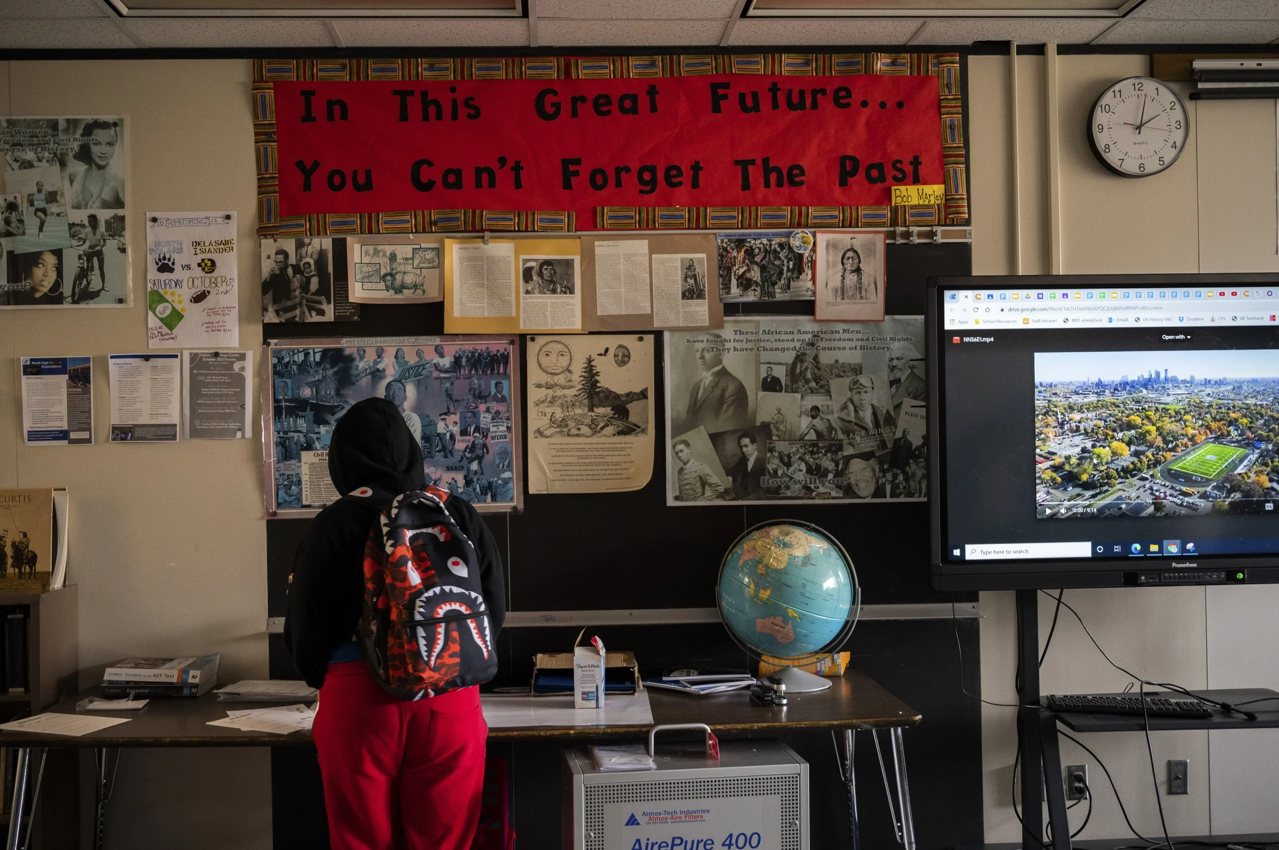 Student with a backpack looking at a classroom bulletin board with historical images, a red banner with the quote, 'In This Great Future... You Can't Forget The Past,' and a world globe on the desk.