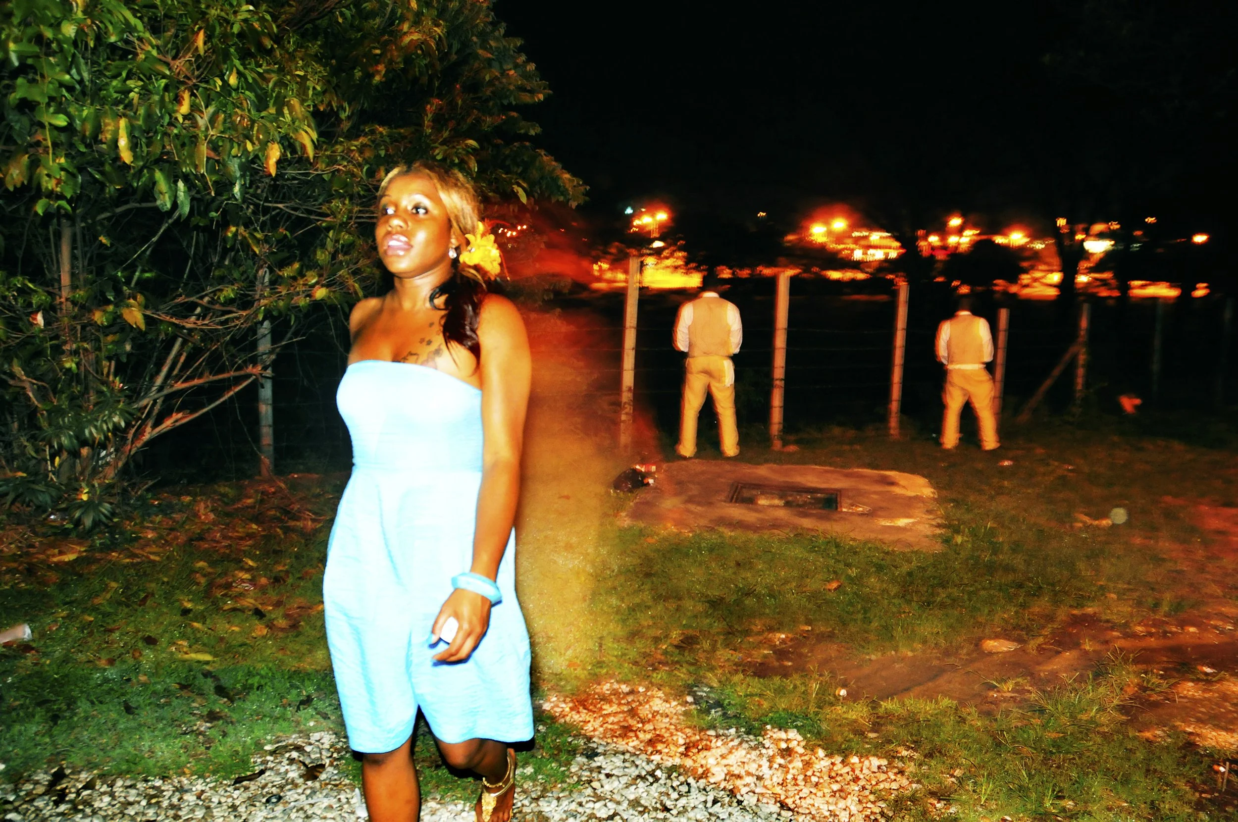 A woman in a blue dress with a flower in her hair standing outdoors at night, with two men in white shirts and yellow vests looking over a fence in the background, illuminated by orange lights.