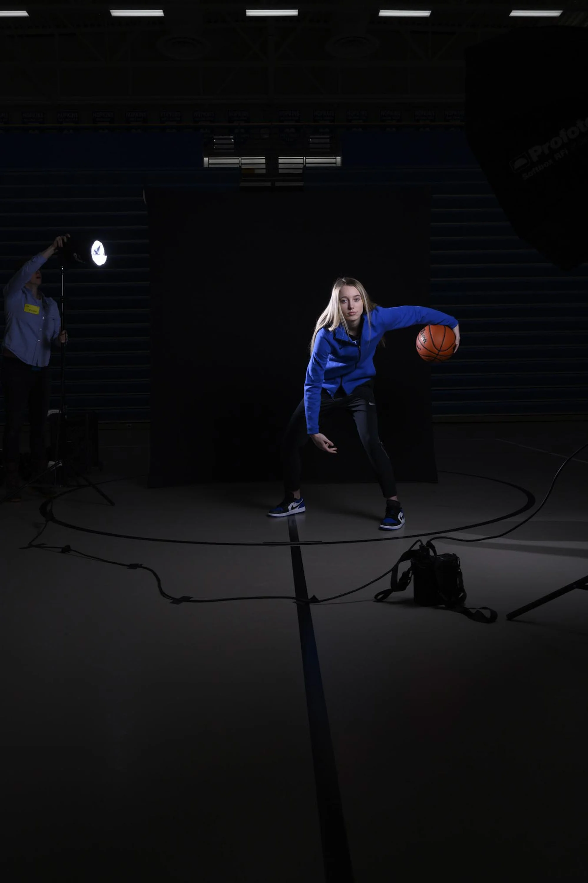 A young woman in a blue athletic jacket dribbling a basketball on a dark indoor court during a professional photo shoot, with studio lighting and equipment visible.