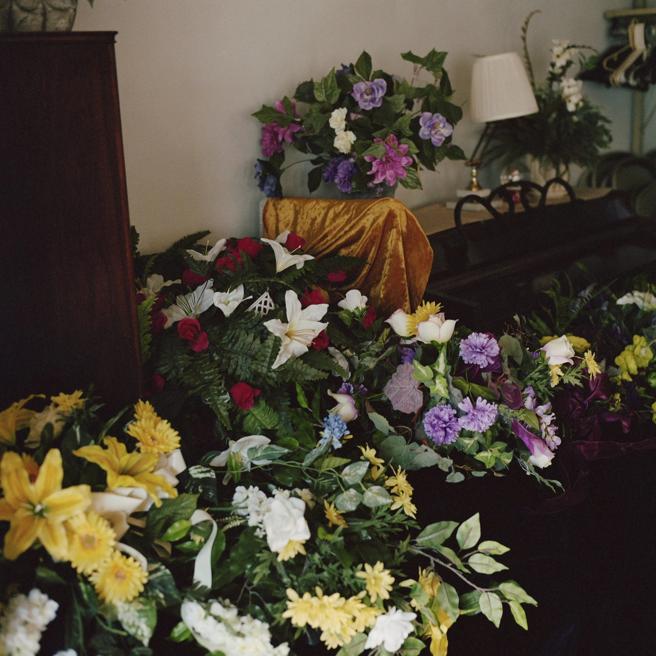 An indoor scene with various colorful flower arrangements, including purple, white, yellow, and red flowers, set against a background that includes a side table with a lamp, a coat rack, and a wall. There is a piece of yellow cloth covering part of t