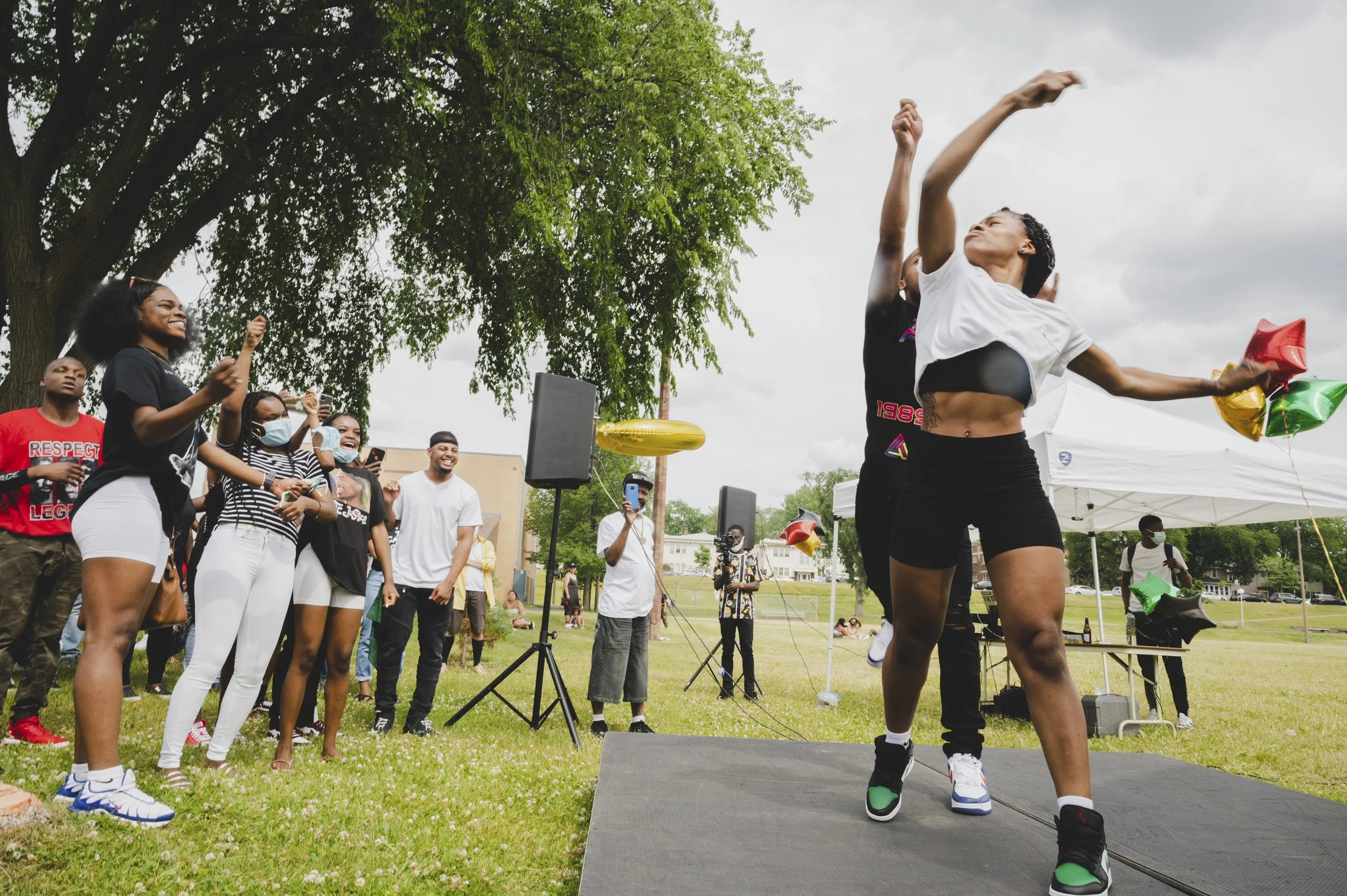 A group of people at an outdoor event, with some smiling and taking photos, while a woman in the foreground is dancing or performing on a small stage holding balloons, with a tree and cloudy sky in the background.