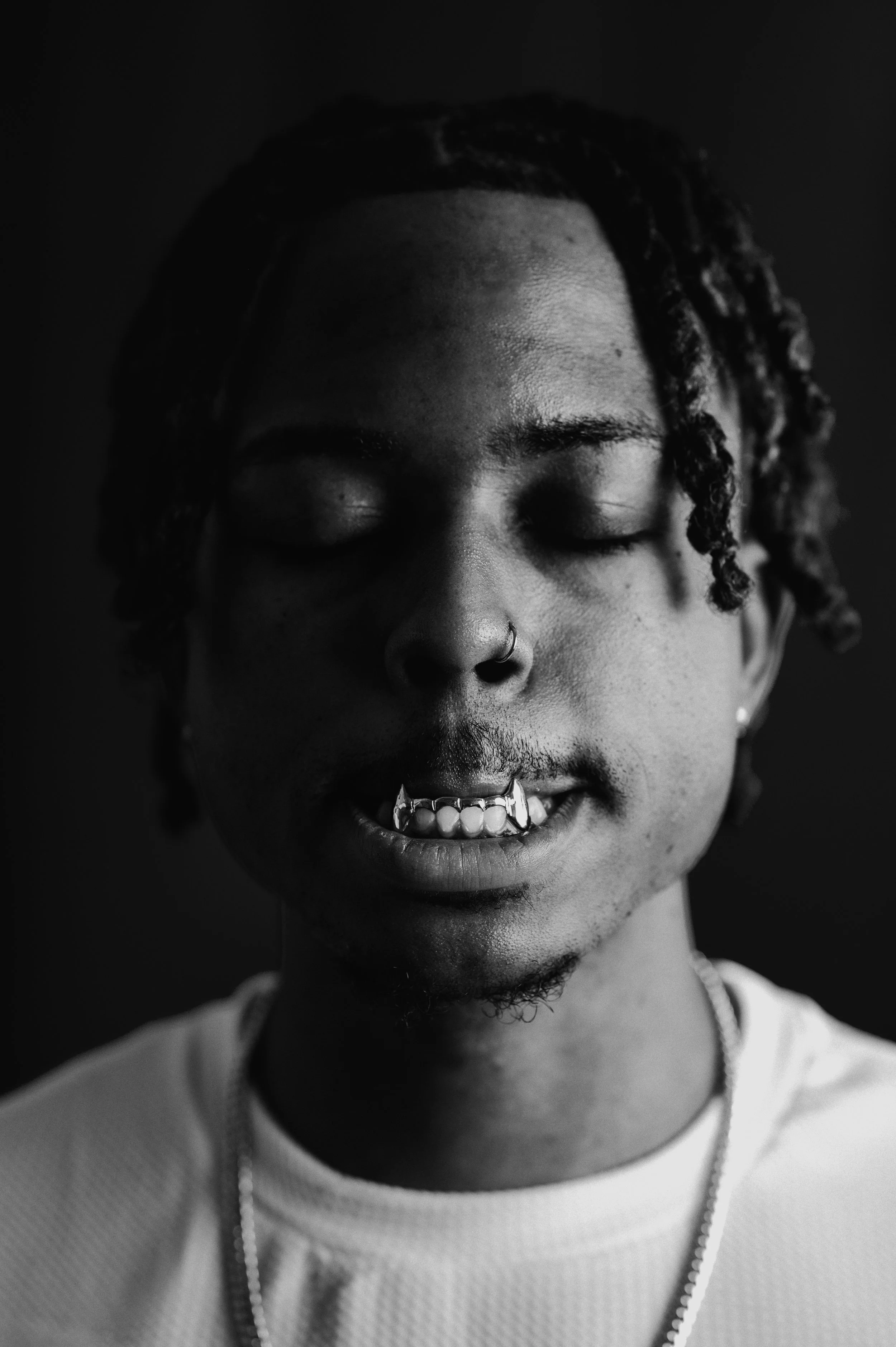 Close-up black and white photo of a young person with dreadlocks, closed eyes, nose piercing, and a chain necklace, smiling slightly.