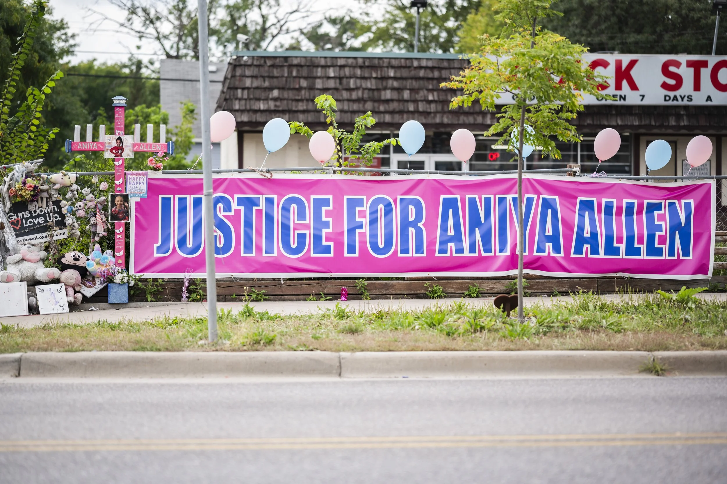 A large pink banner with blue text that reads 'JUSTICE FOR ANYA ALLEN' hangs outside a building. The area around the banner has balloons, stuffed animals, flowers, and photos, indicating a memorial or vigil for Anya Allen.