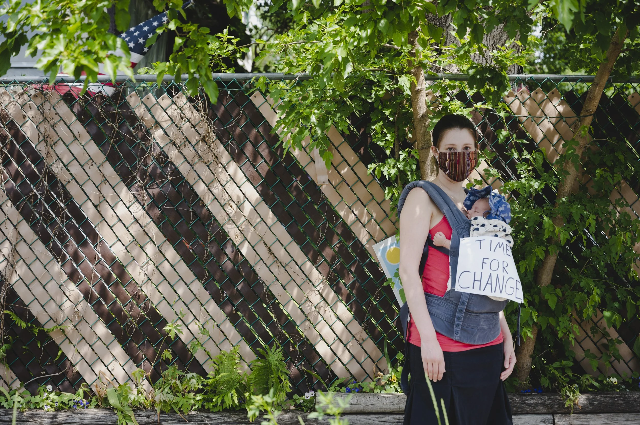 A person standing outdoors with a baby in a carrier, wearing a face mask, holding a sign that reads 'Time for Change,' in front of a garden fence and green trees.