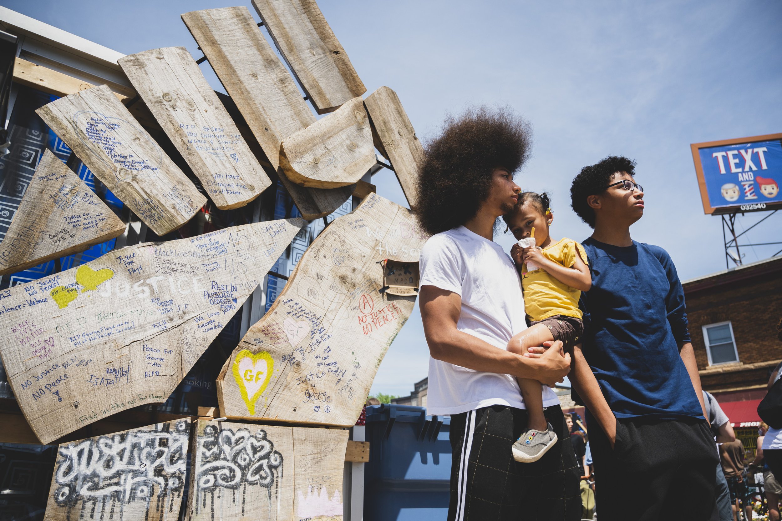 Two young adults, a man and a woman, standing together outdoors with a little girl. The man is holding the girl, who is eating a popsicle, while the woman stands beside them. In the background, there is a display of wooden planks with handwritten mes