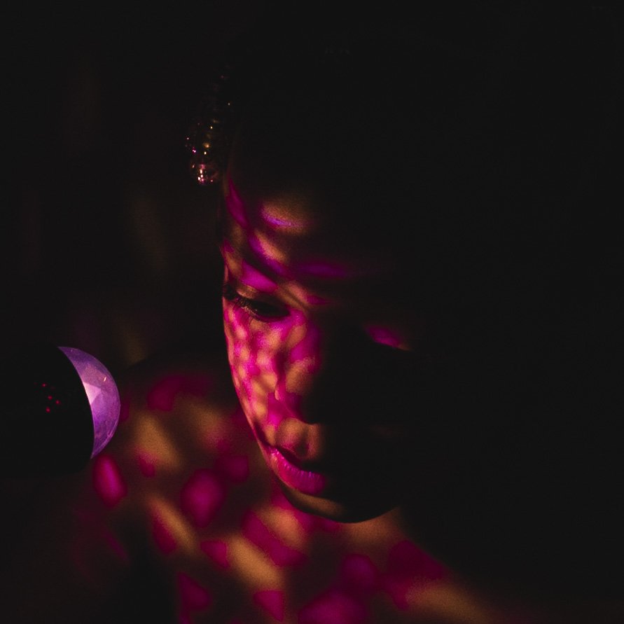 A child's face is illuminated by pink and purple light, with a reflective pattern projected onto their skin, in a dark environment.