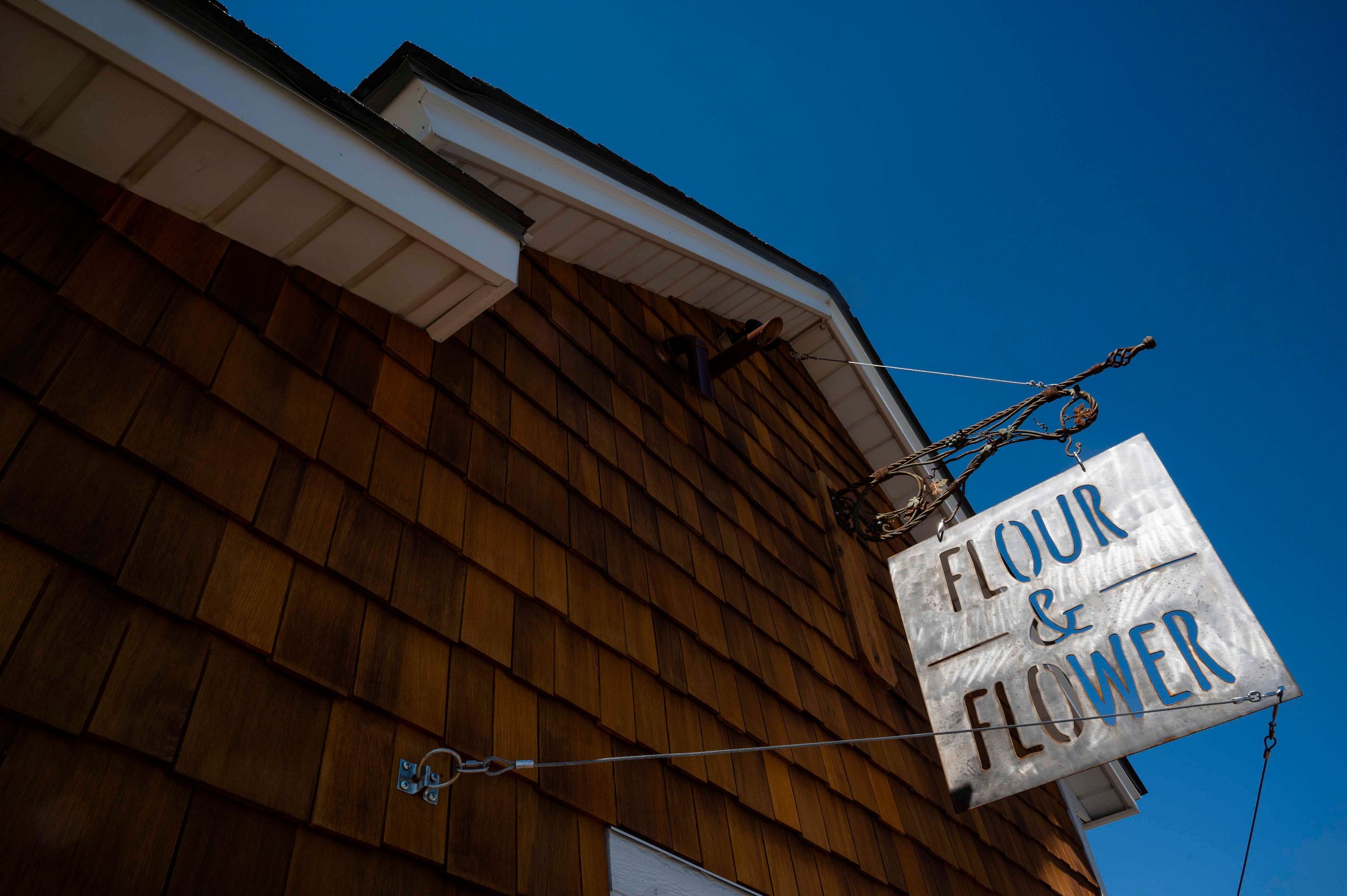 A metal sign reading 'FLOUR & FLOWER' is hanging outside a building with brown shingles, attached by metal chains to a decorative iron bracket against a blue sky.