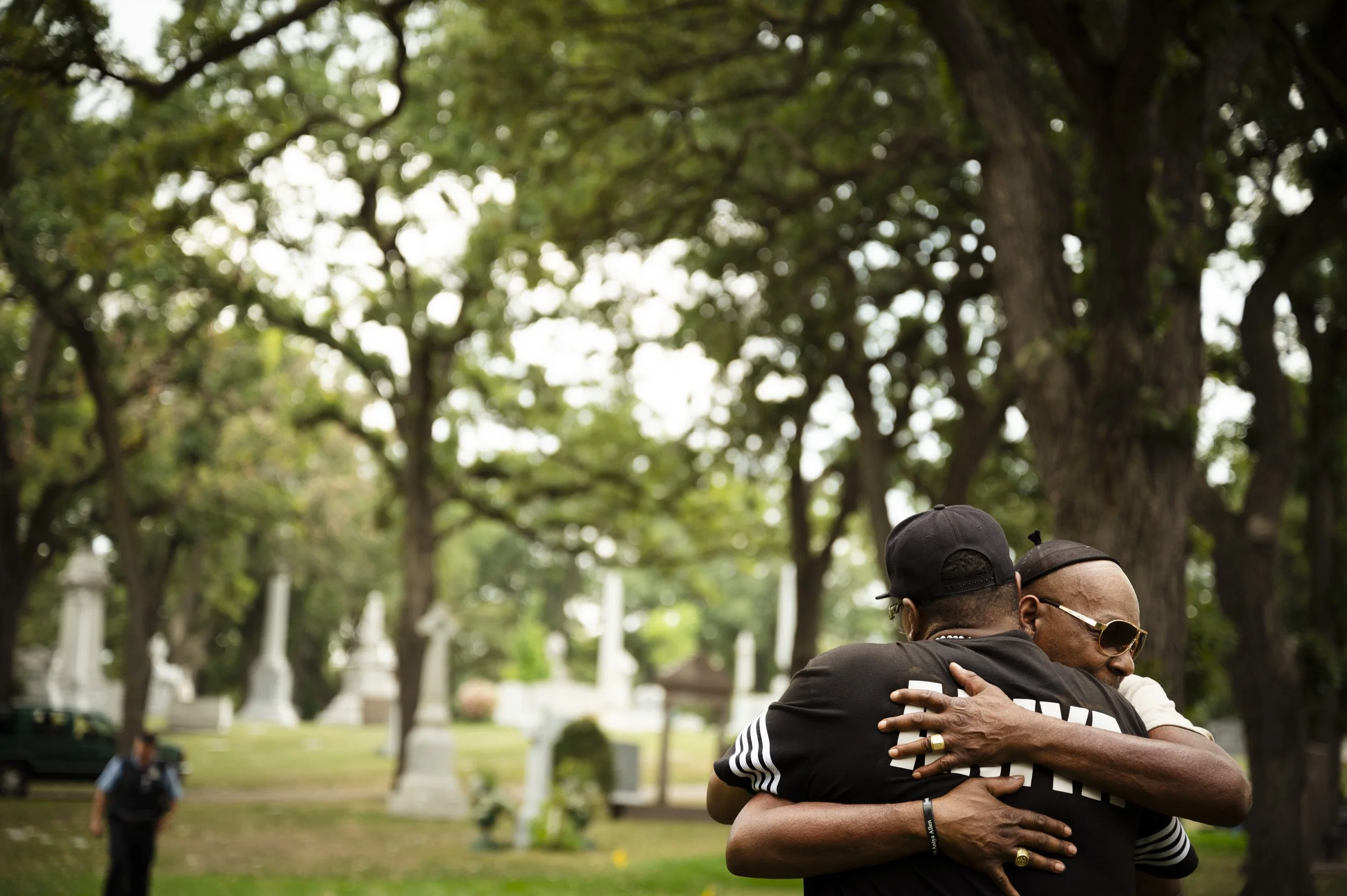 Two men hugging tightly outdoors in a park-like setting with large trees and tombstones in the background.