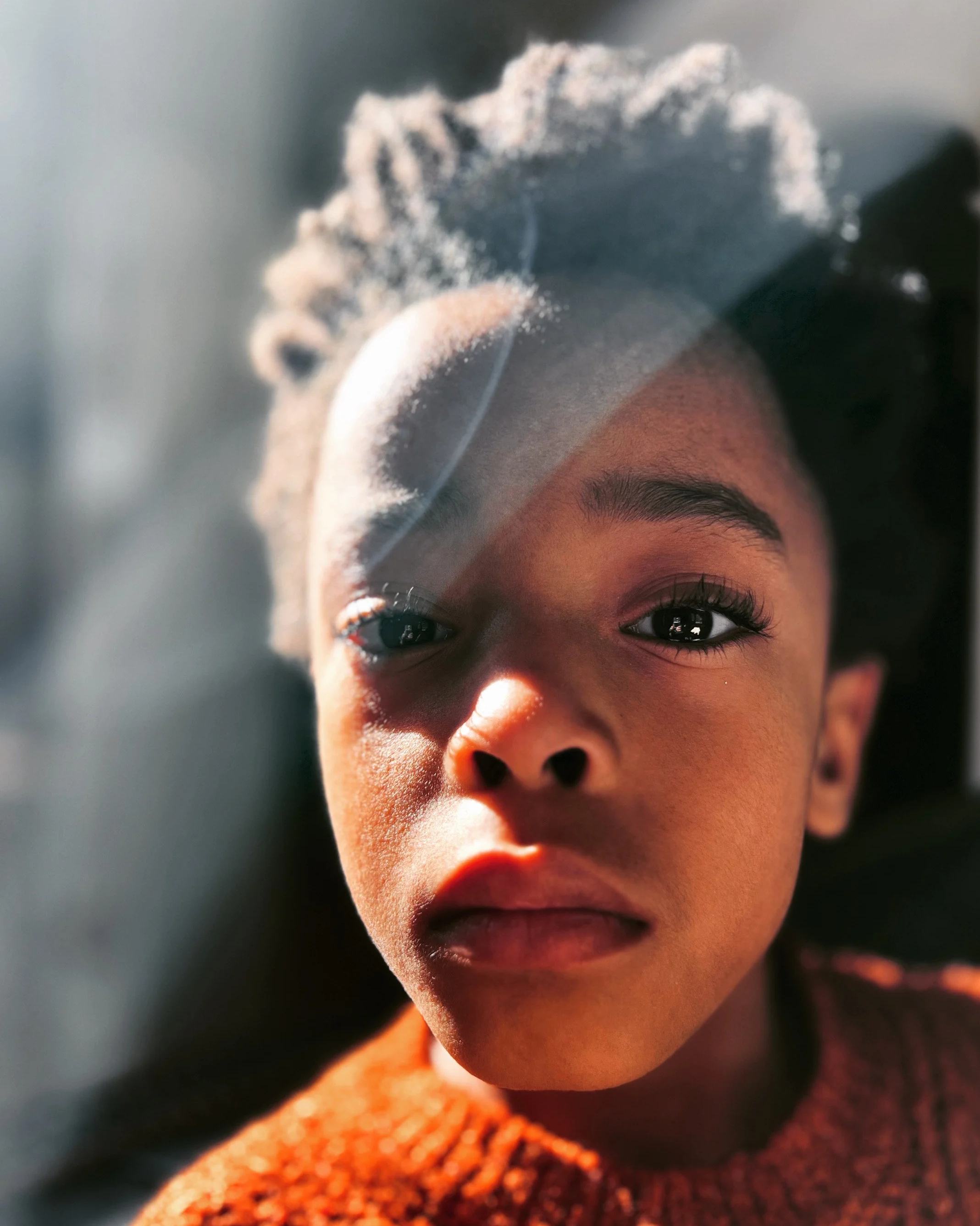 Close-up of a Black child's face with sunlight and a window reflection, wearing an orange sweater.