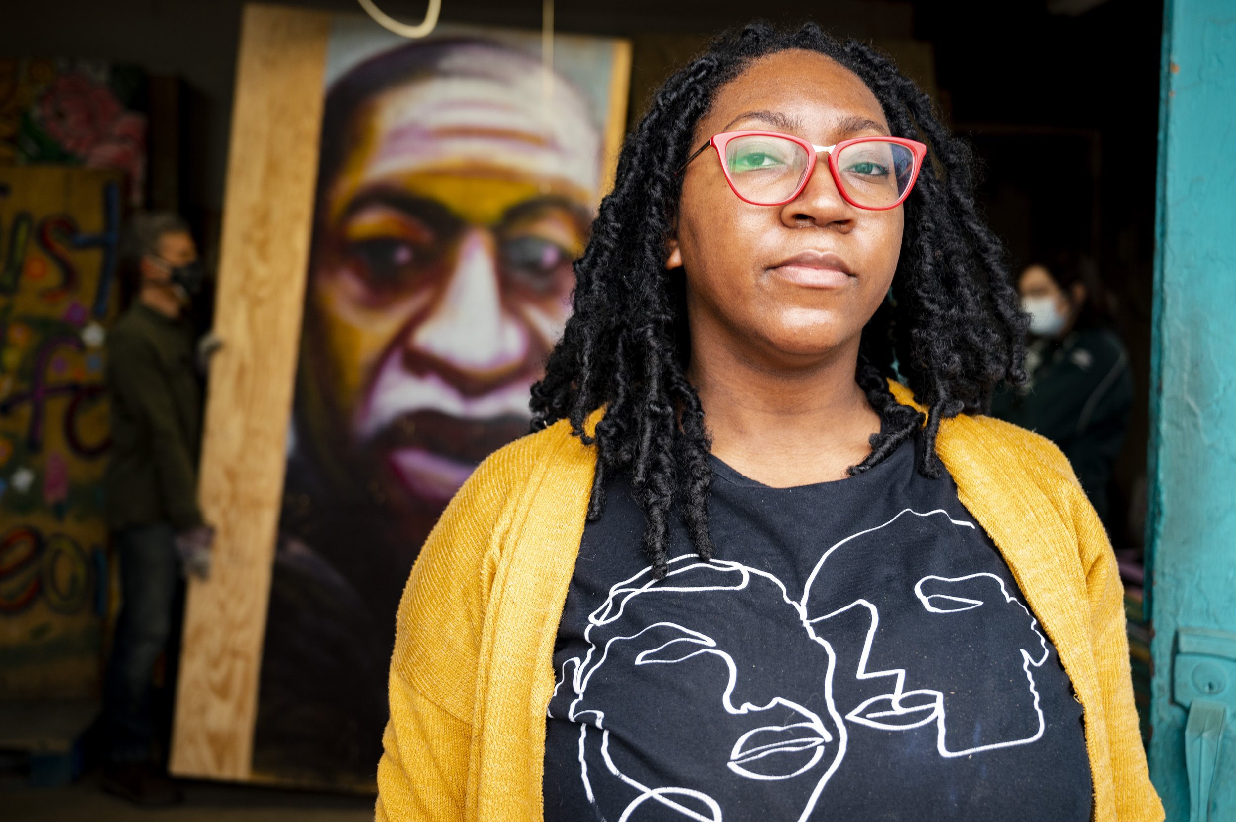 Portrait of a woman with black dreadlocks wearing red glasses, a black t-shirt with abstract line art of faces, and a yellow cardigan in front of a large colorful portrait of a person with glasses.