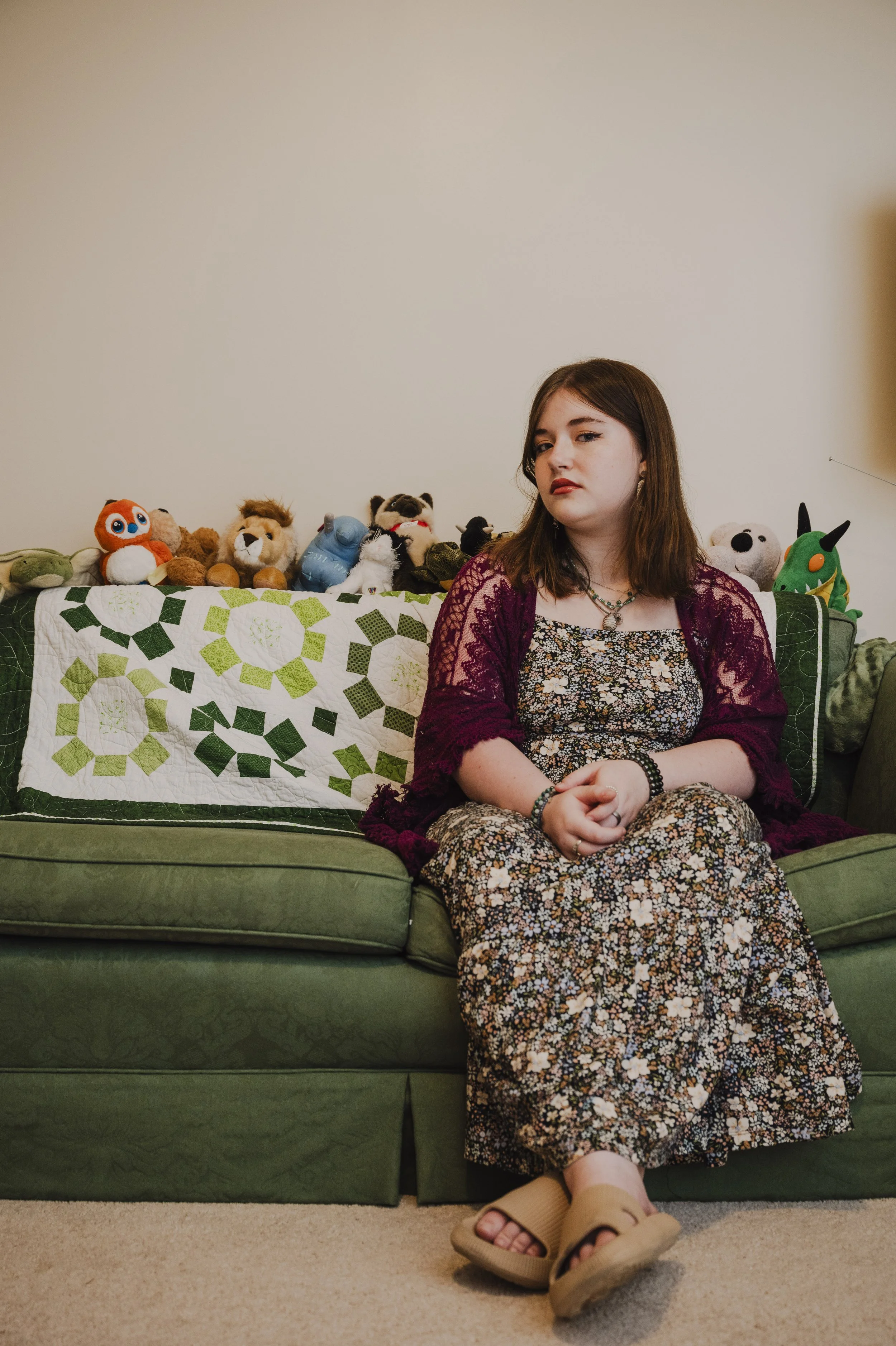 A young woman sitting on a green couch with a floral-patterned skirt, wearing a maroon lace cardigan and sandals, with stuffed animals arranged on the back of the couch, including a fox, lion, raccoon, panda, and a green plush toy.
