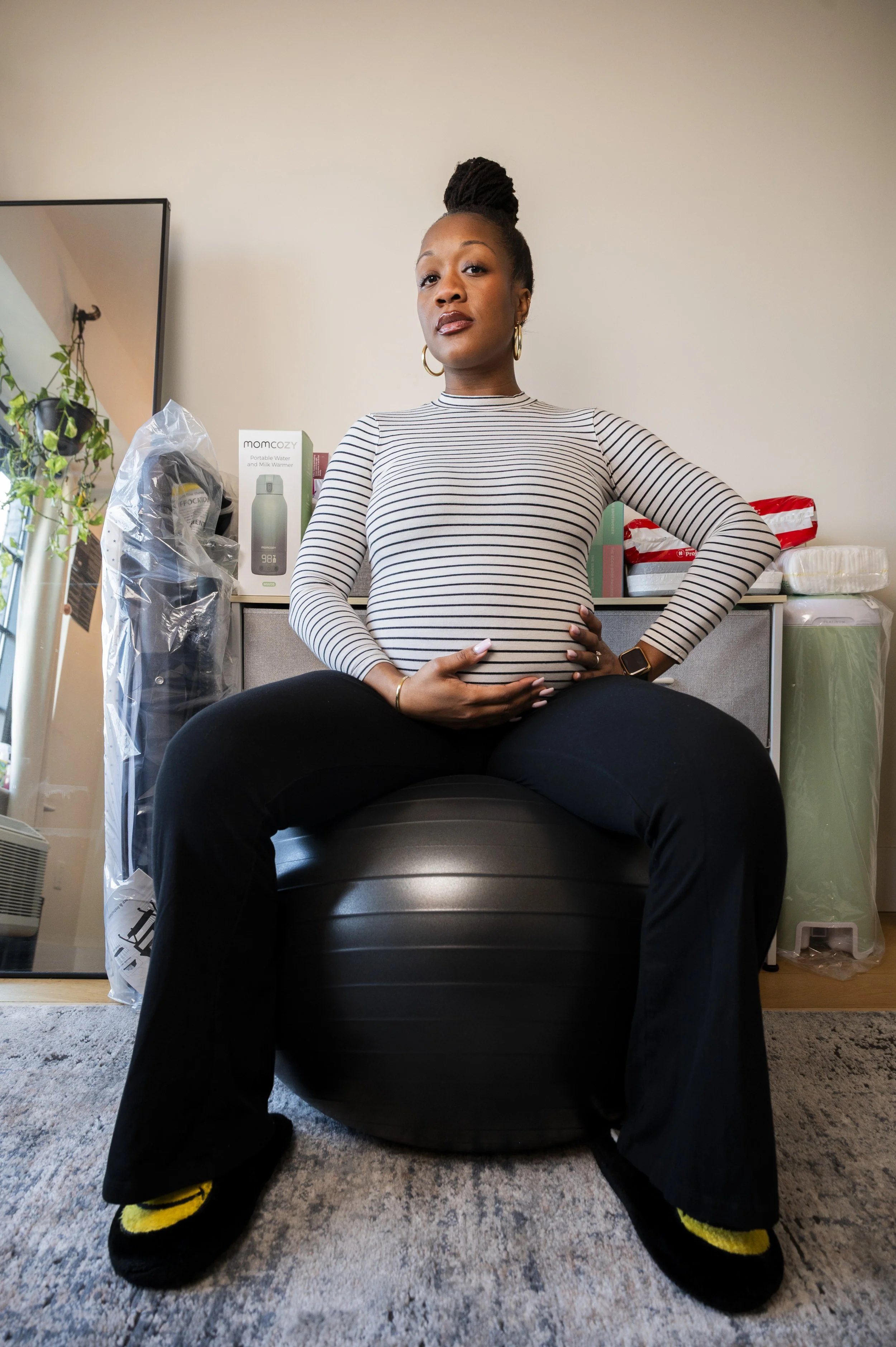 A pregnant black woman sitting on an exercise ball indoors with one hand on her pregnant belly and the other on her hip, looking confidently at the camera.