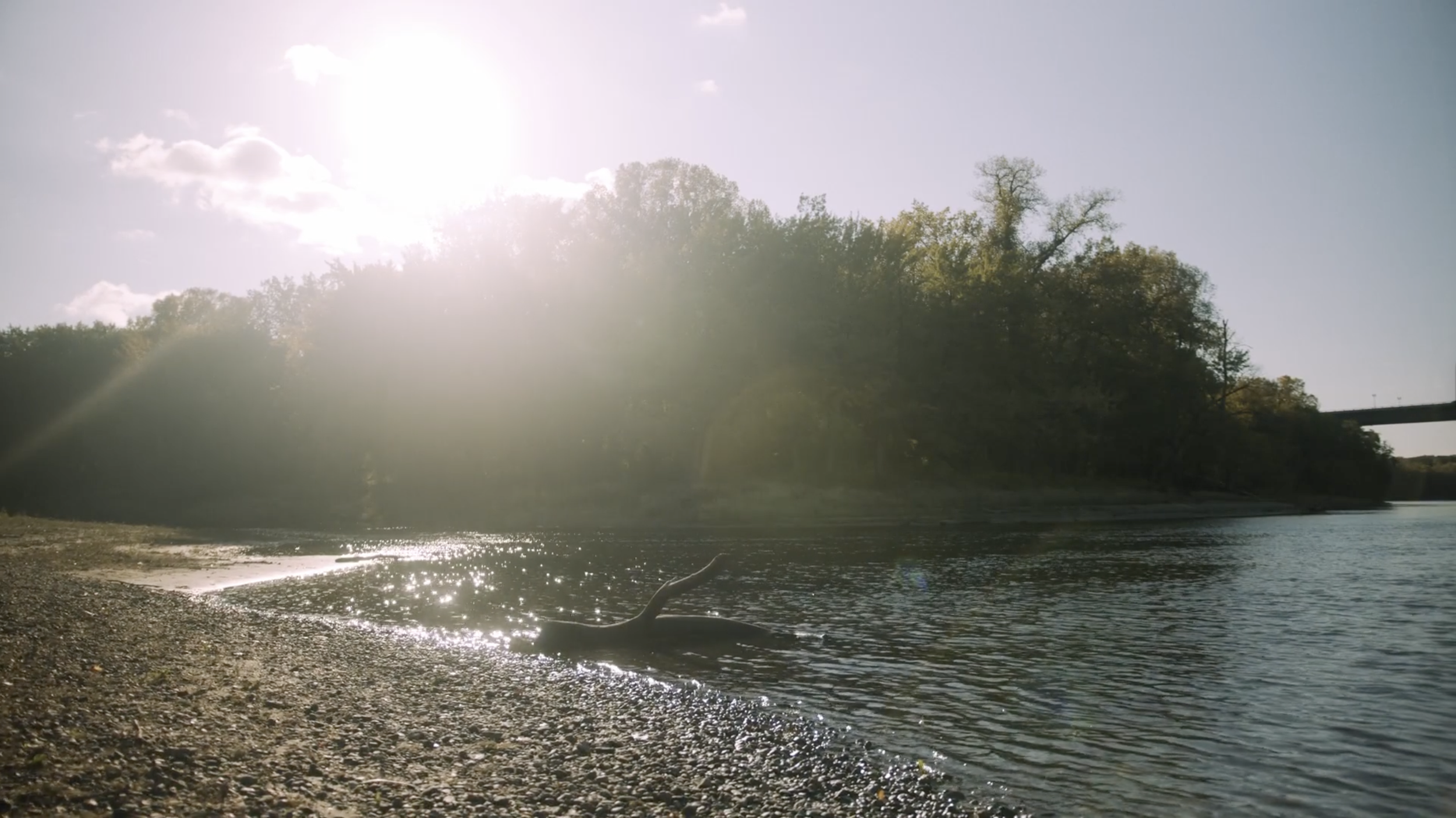 A riverside scene in the late afternoon with the sun shining brightly, casting reflections on the water. The riverbank is covered with small pebbles, and there is a branch partially submerged in the water. Trees line the opposite riverbank with a bridge visible in the distance to the right.