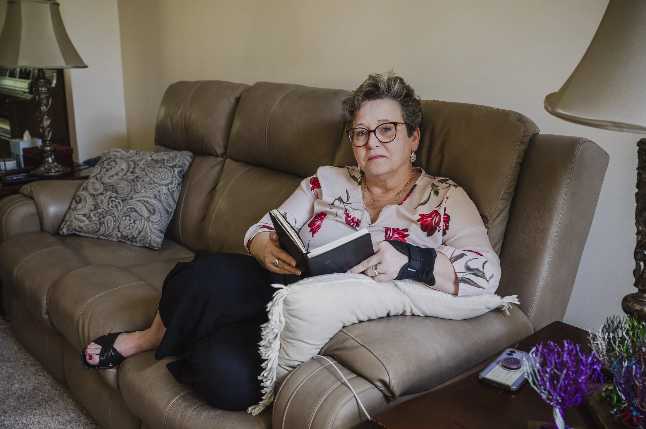 An older woman with short gray hair, glasses, and earrings sitting on a beige leather couch in a living room. She is wearing a floral blouse, holding an open notebook, and looking at the camera. There are lamps on side tables and decorative pillows o
