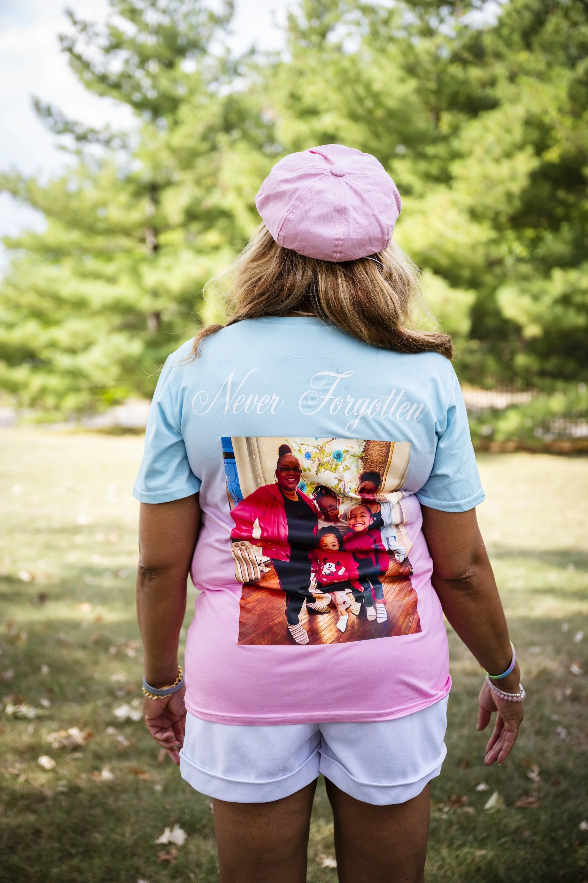 Back of a woman wearing a pink beret and a blue shirt with a large family photo and the words "Never Forgotten," standing outdoors in a park with green trees.