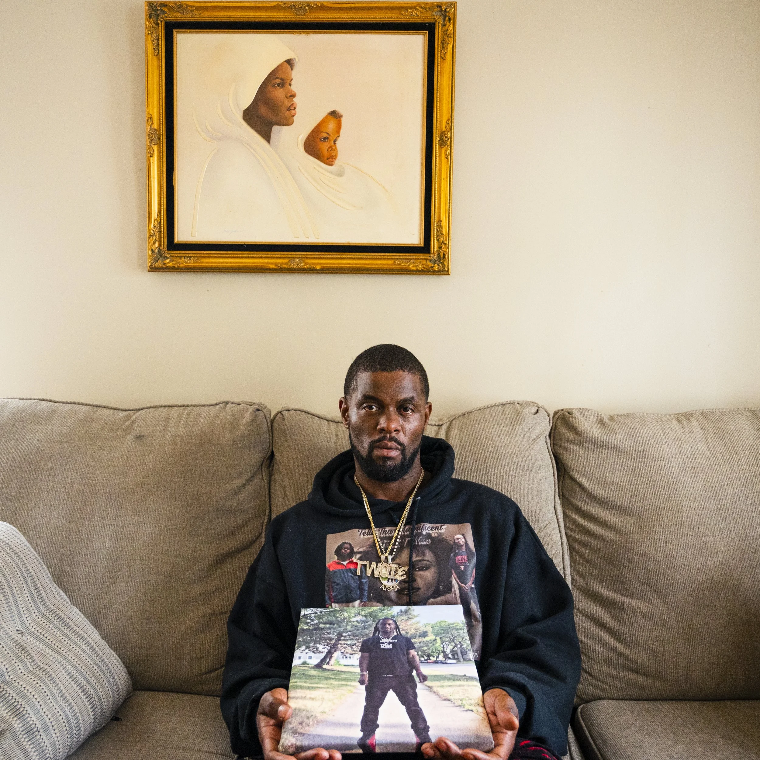A man sitting on a beige couch, holding a photo of himself standing outdoors in a park. He wears a black hoodie with graphic designs and jewelry, looking directly at the camera with a serious expression. Behind him is a framed painting of two women in white garments on a plain wall.