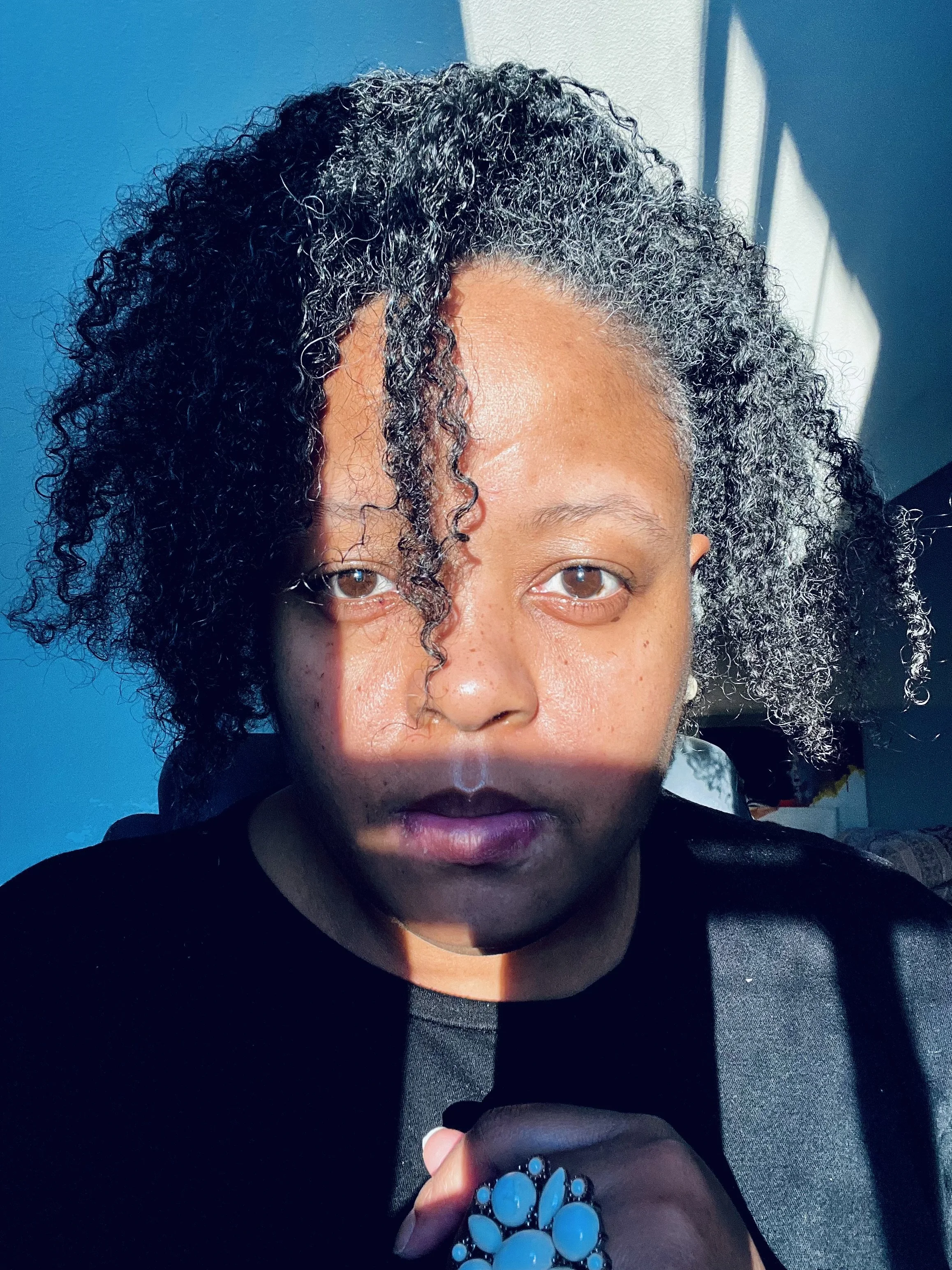 A Black woman with curly hair in sunlight, revealing one side of her face in shadow, wearing a large flower ring.