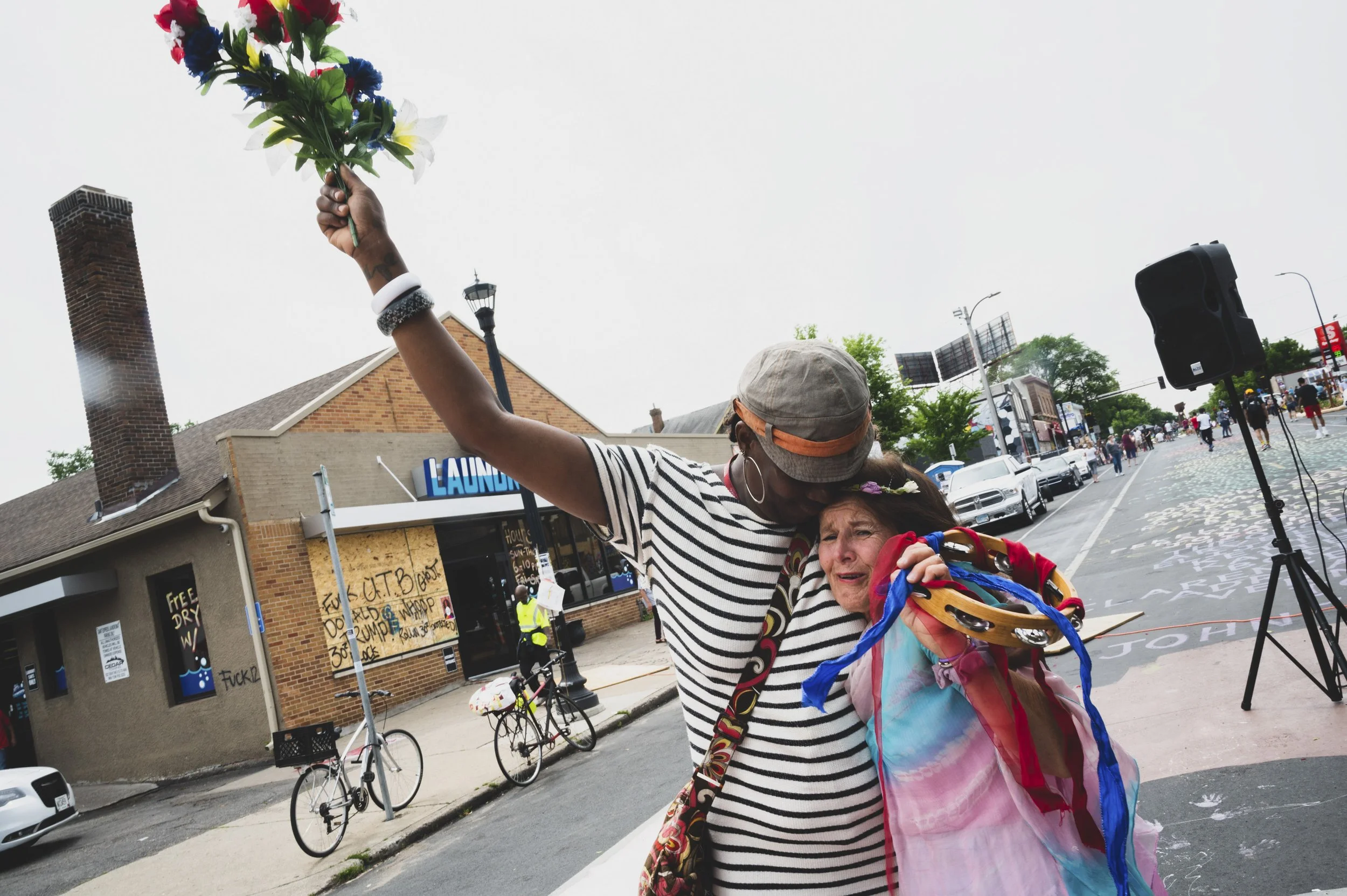 A person holding a bouquet of colorful flowers above an elderly woman in a decorated wheelchair during an outdoor event on a city street.