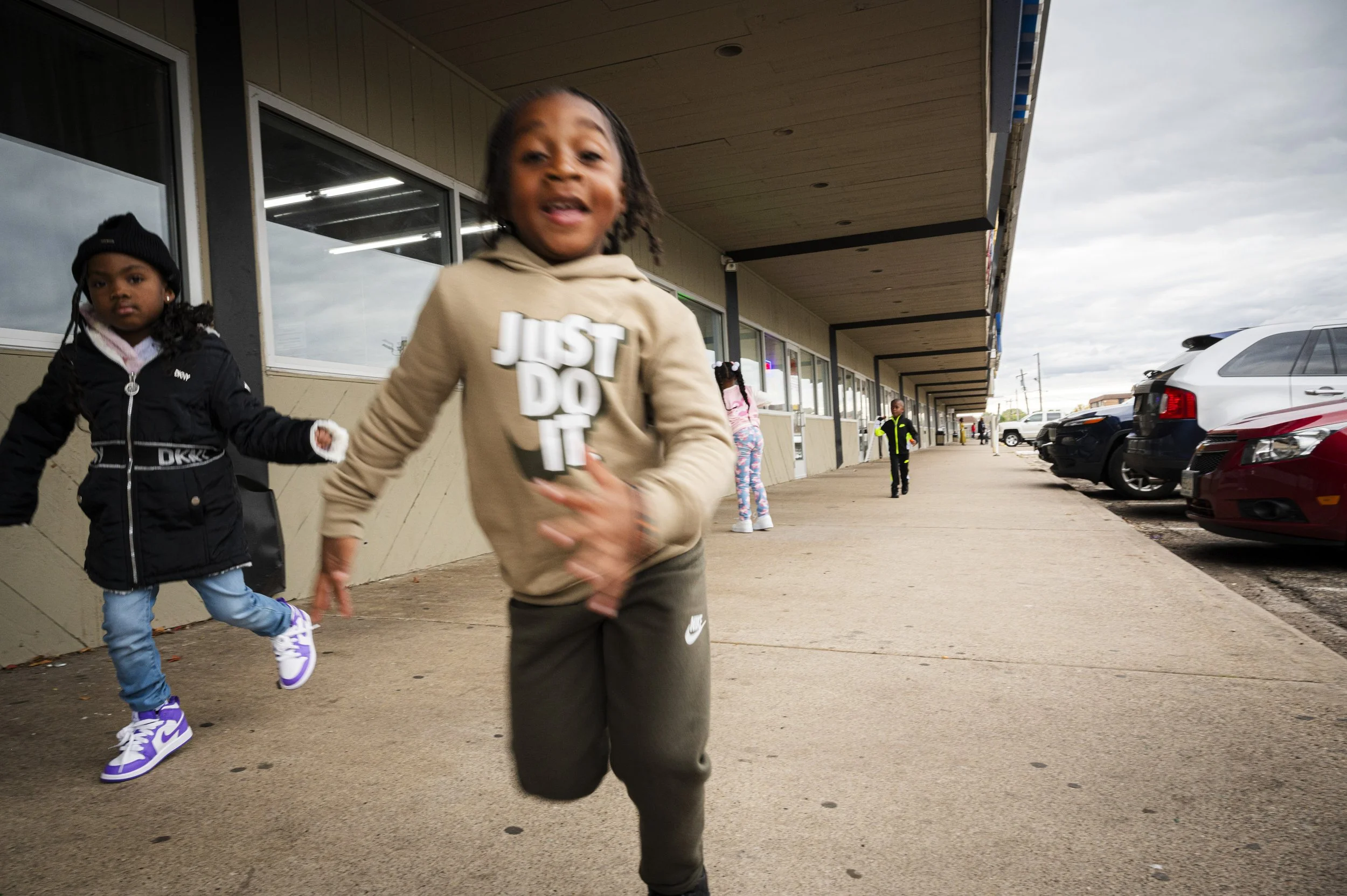 Two children running outside a shopping center, with several parked cars and a cloudy sky in the background.