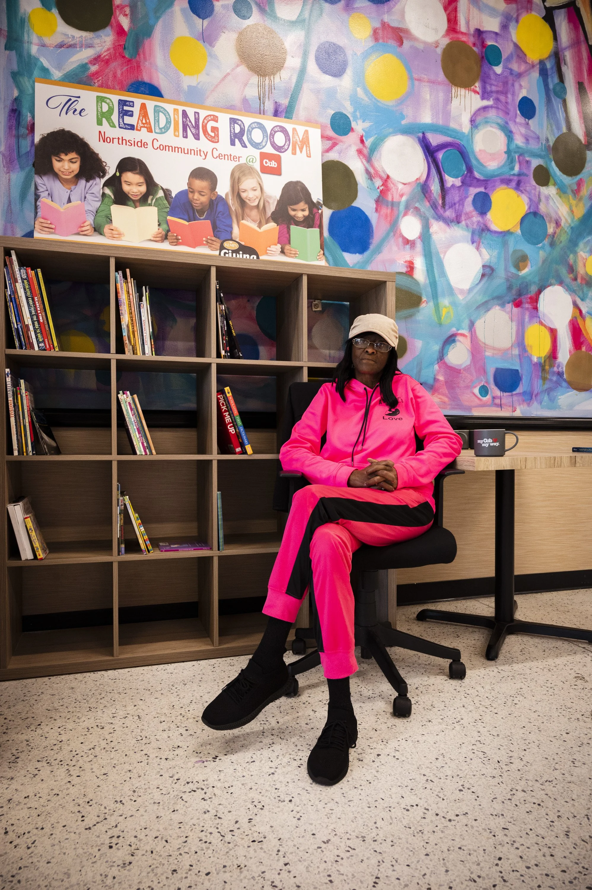 A woman in a bright pink tracksuit sits in a black office chair in front of a colorful abstract mural and a bookshelf filled with books. She is wearing glasses and a light beige cap.