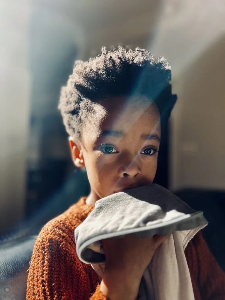 A young child with short, curly hair holding a cloth up to their face, with sunlight shining through a window illuminating their face and hair.