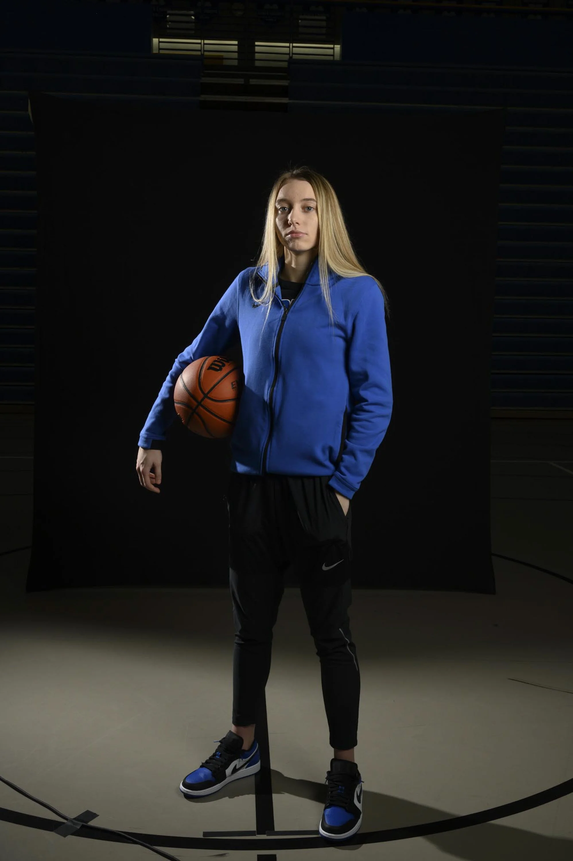 Young woman in blue sports jacket holding basketball on a gym basketball court.