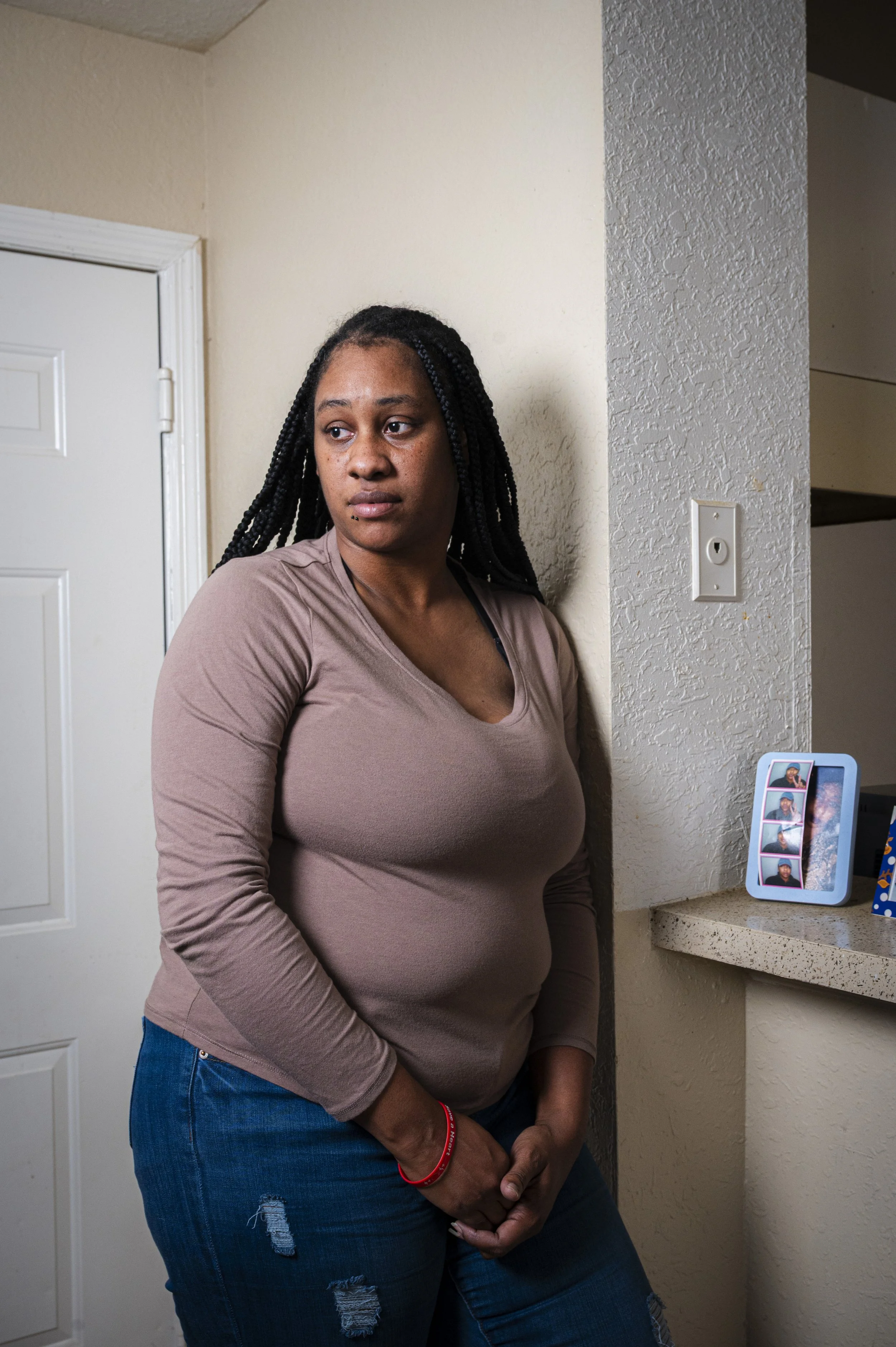 A woman with black braided hair standing in a room with beige walls, looking away from the camera with a serious expression, wearing a light brown long sleeve shirt and ripped jeans.