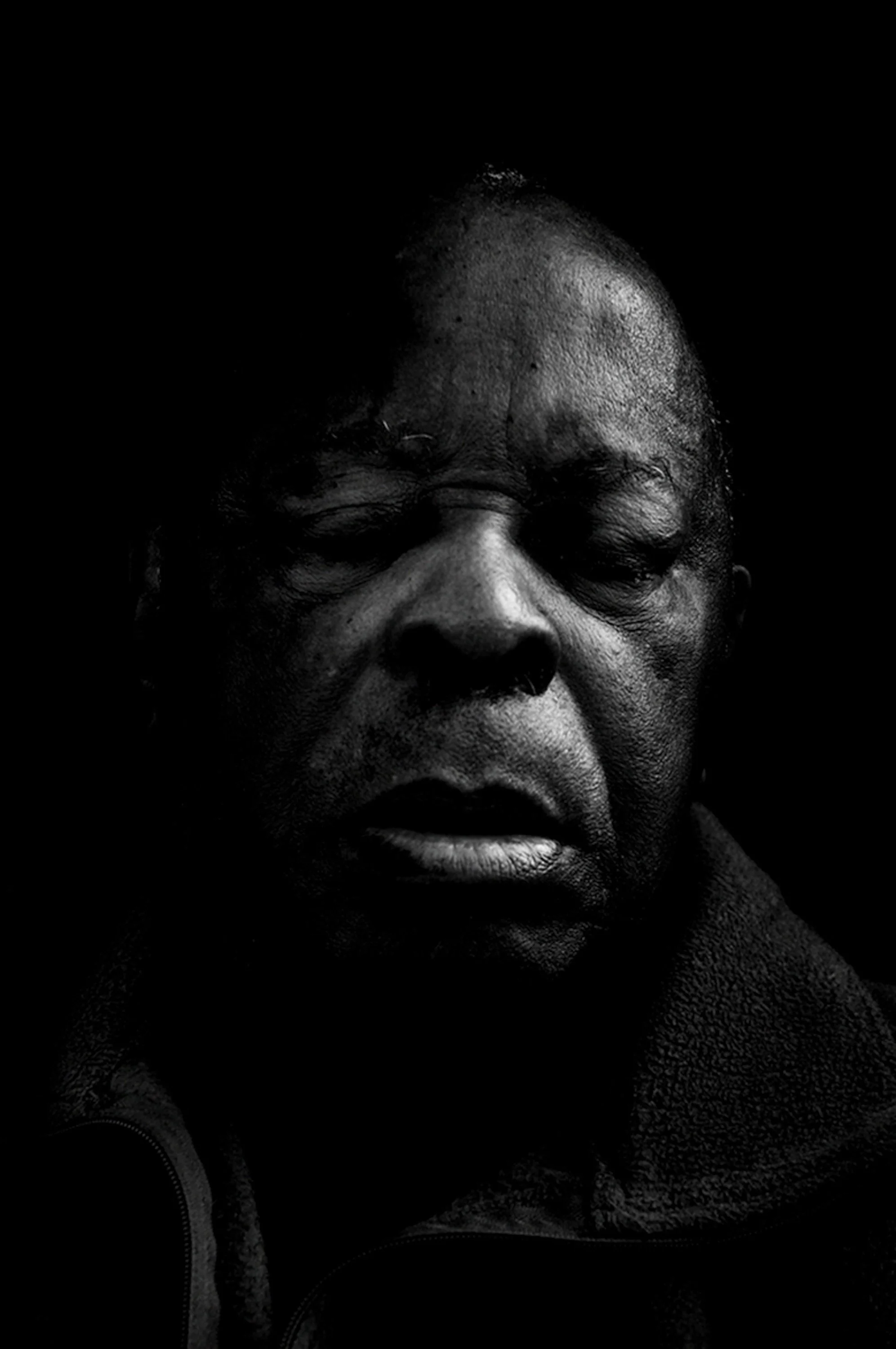 Black and white close-up portrait of an older Black man with eyes closed, showing detailed facial features and a textured coat collar. He is tired and has closed his eyes to get some rest.