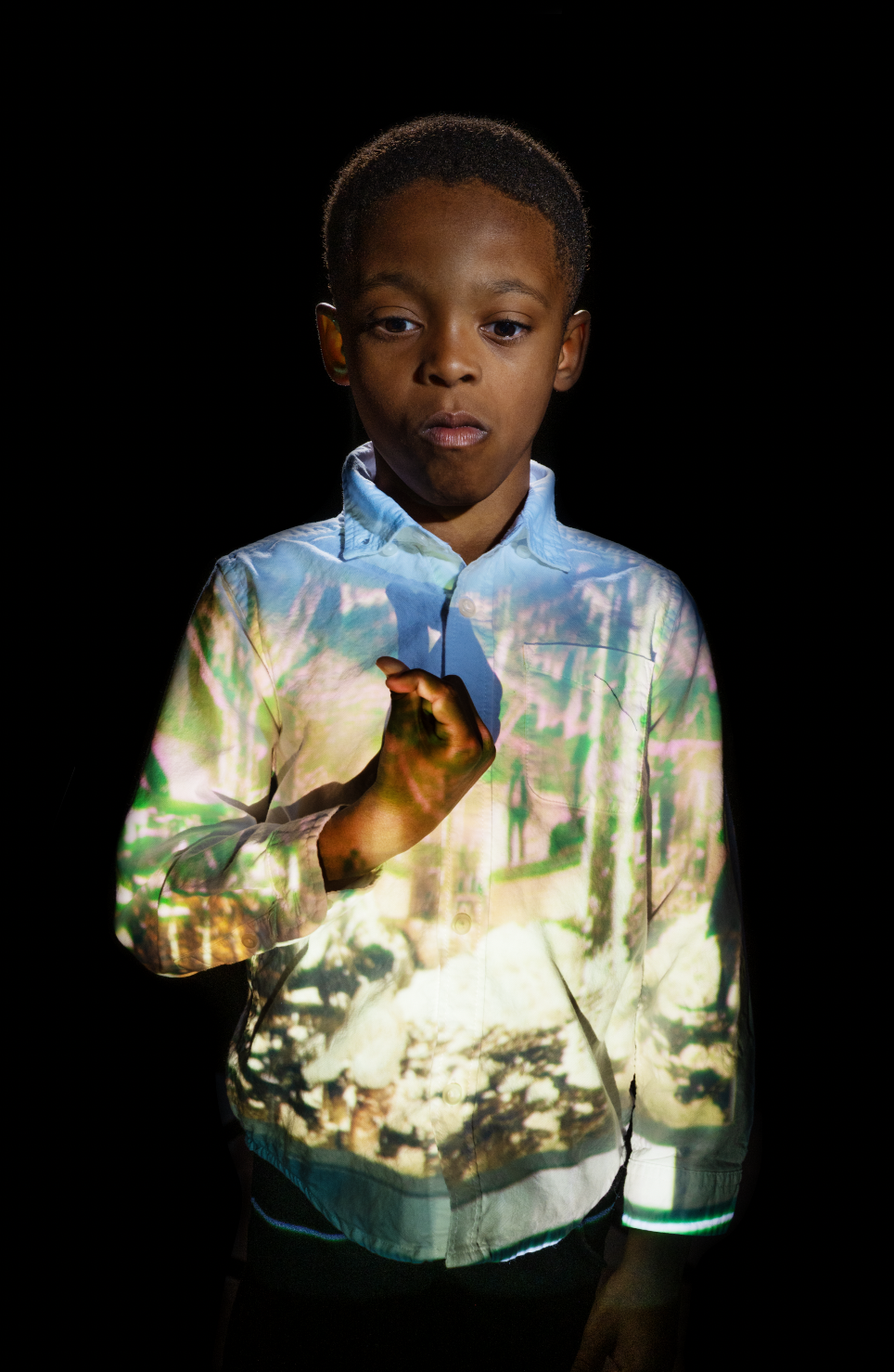 A young boy standing in a dark room with a projection of a colorful, scenic landscape on his shirt and face.