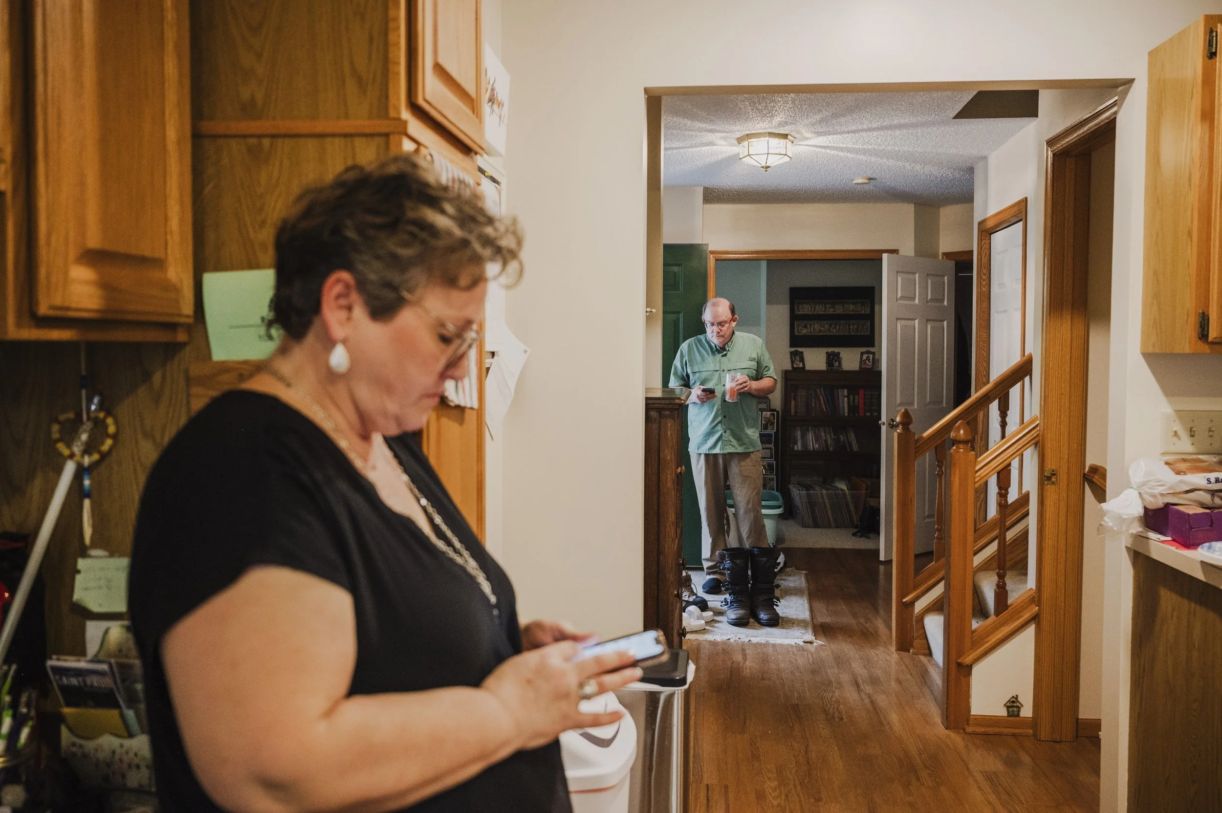 A woman in the foreground using her phone, partially visible, with a man in the background holding a drink and looking at his phone inside a home with wooden cabinets and a hallway visible.