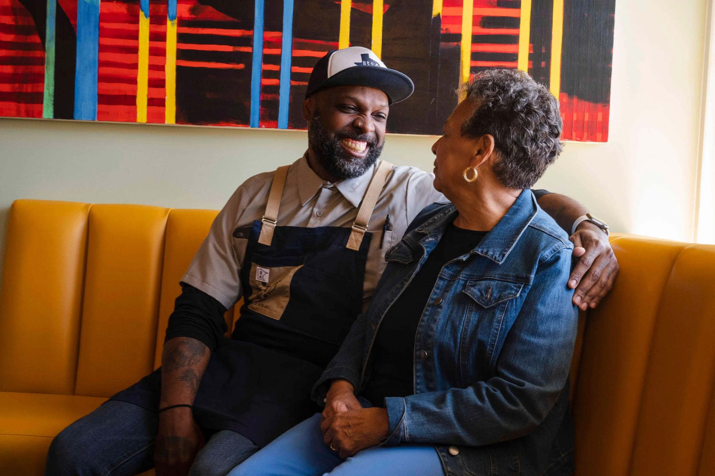 A man and woman sitting on a yellow couch, smiling, looking at each other, with a colorful abstract painting on the wall behind them.