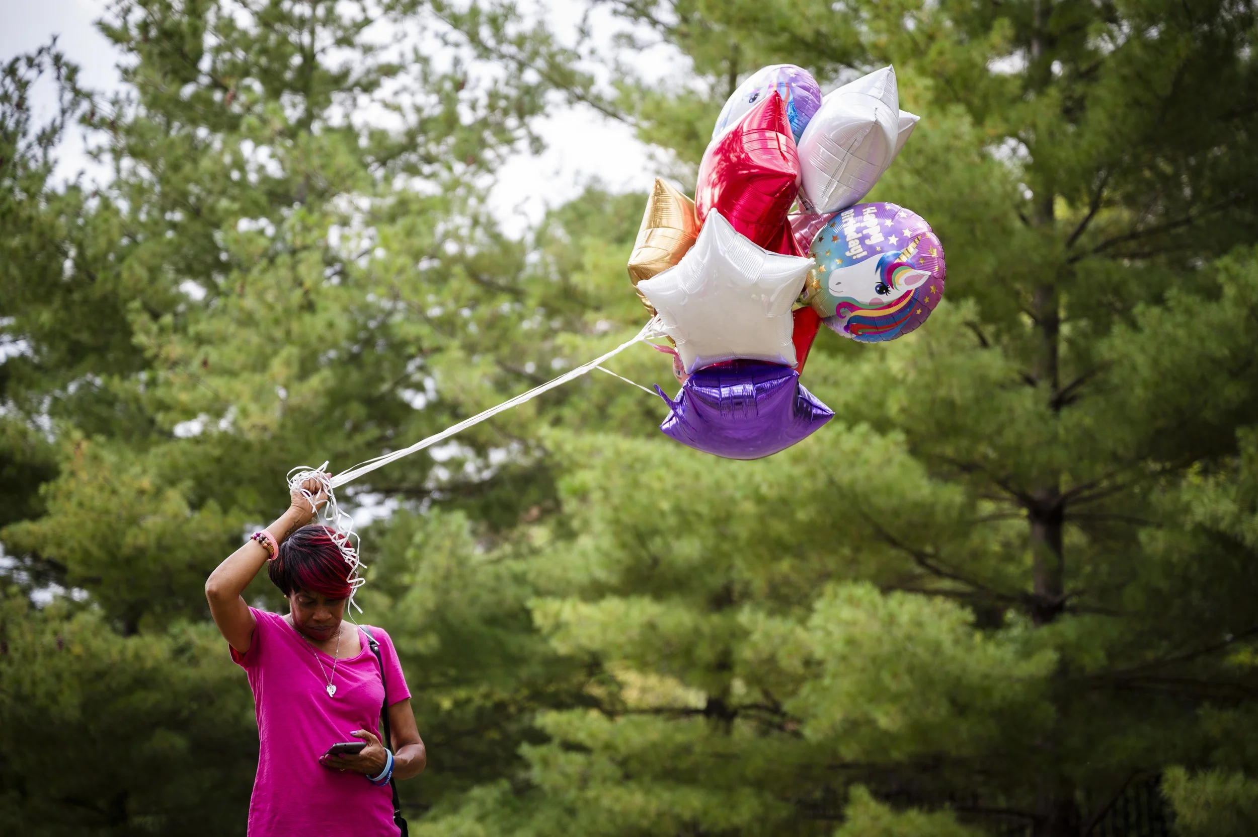 A woman wearing a pink shirt holding a bunch of colorful helium balloons in a park with green trees in the background while looking at her phone.